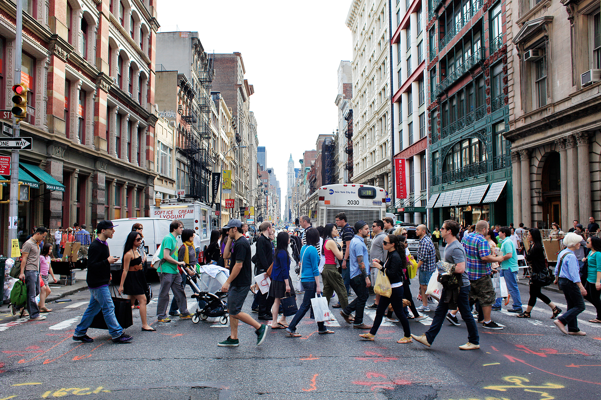 A busy street in SoHo, NYC with numerous people crossing at a crosswalk in different directions. The background features tall buildings, storefronts, and a bus. The sky is clear, and the ground has colorful markings. Photo by Joe Buglewicz/NYC and Company.