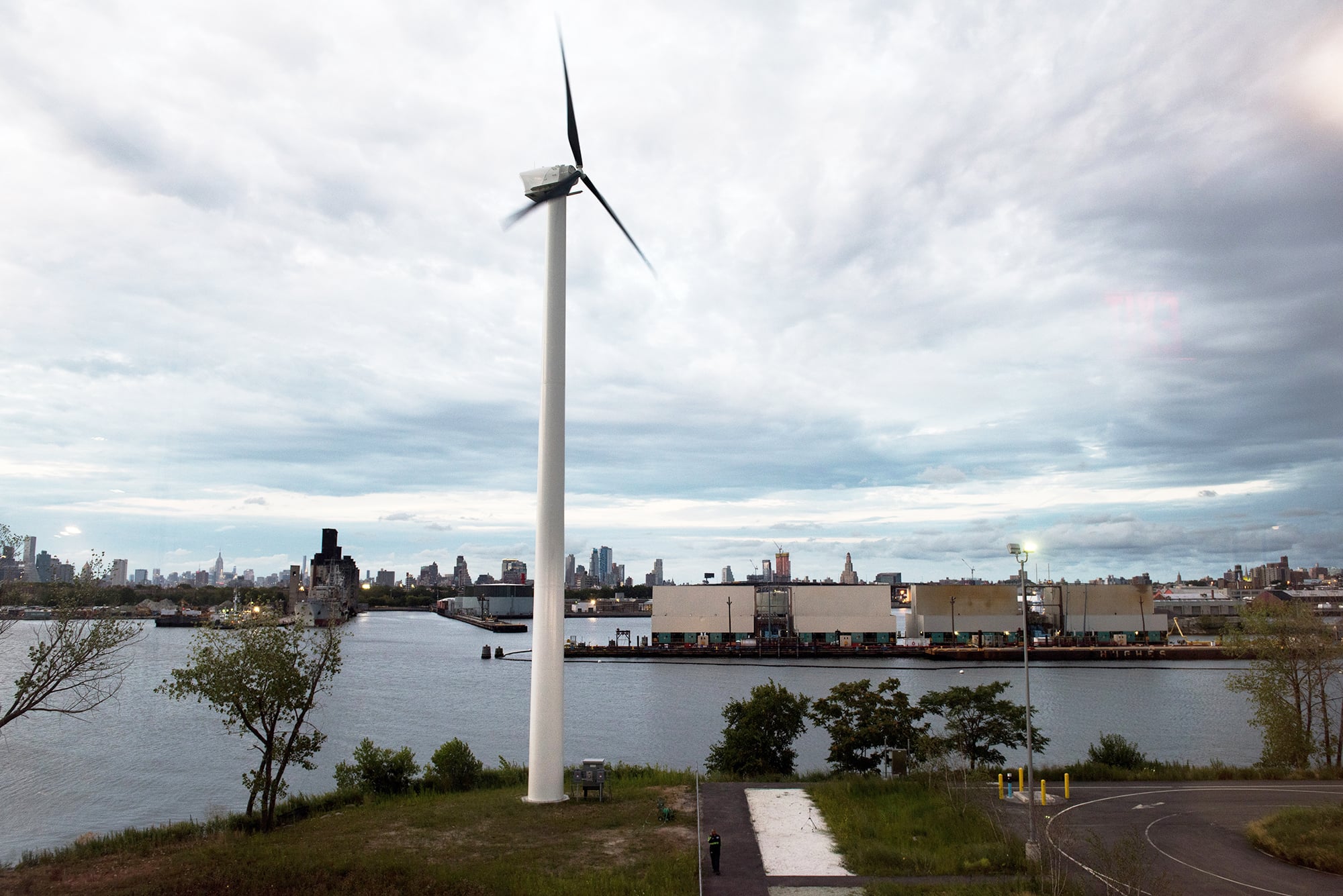 A wind turbine stands near a river, with an industrial area and NYC skyline in the background. The sky is cloudy, and trees and greenery are visible in the foreground.