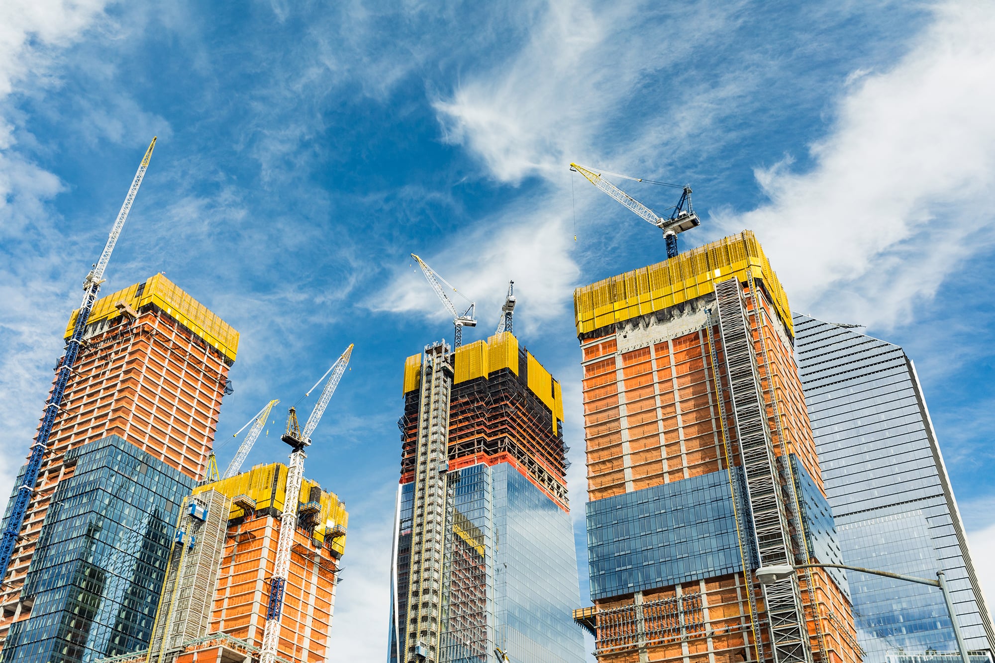 Tall skyscrapers under construction in Hudson Yards with cranes on top against a blue sky with white clouds. The buildings have glass and steel exteriors, with yellow and orange safety netting around upper floors.
