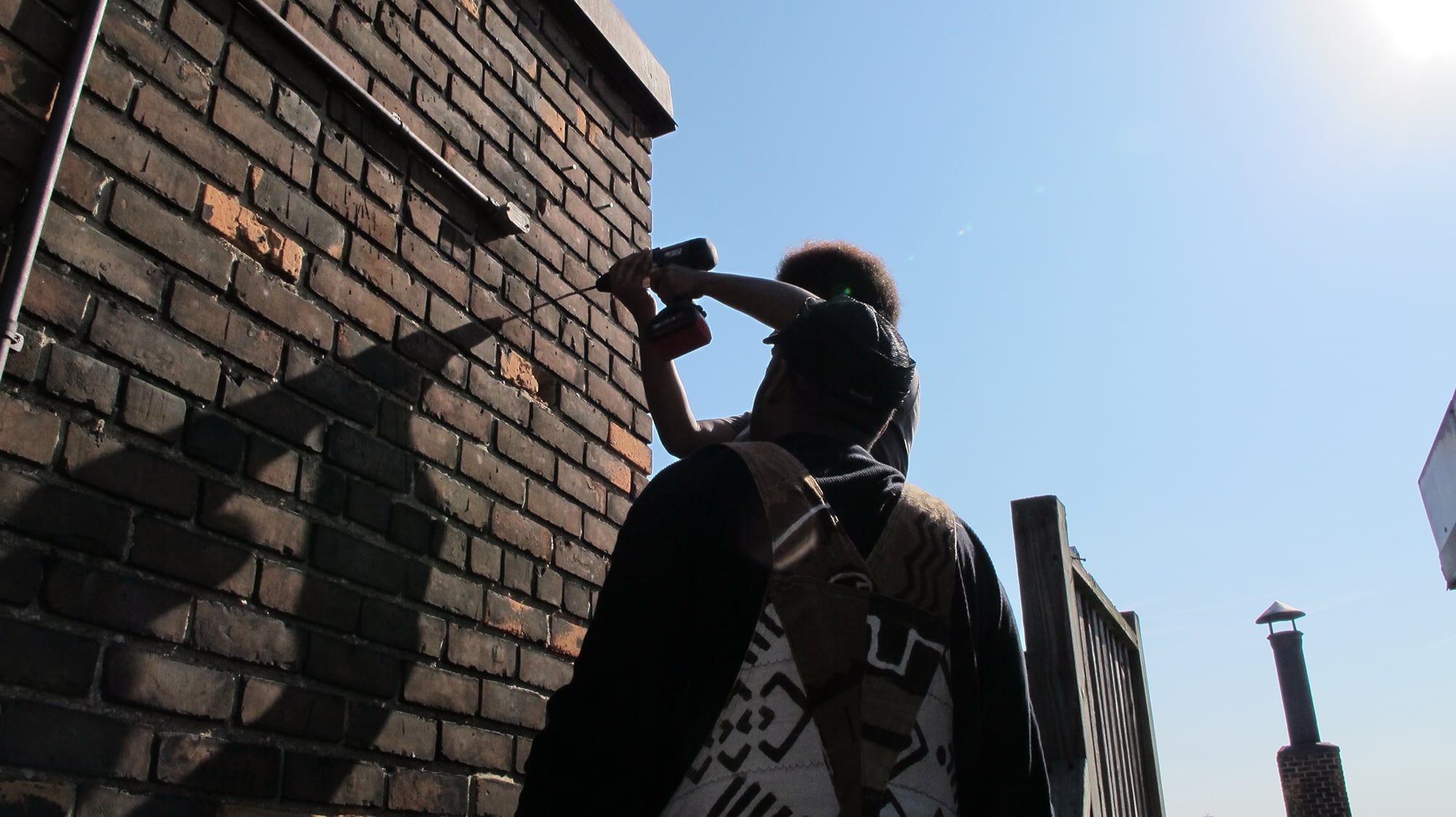 Body image for Rise NYC. Two Detroit Digital Stewards work on a brick wall under a clear blue sky. One is using a power drill, while the other stands beside, wearing a harness. Sunlight creates shadows on the walls surface.
