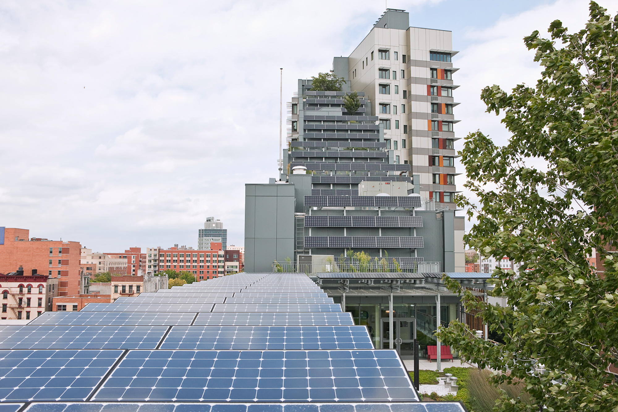 A modern building with multiple balconies covered in greenery, set against a cityscape. In the foreground, solar panels are installed on a roof, reflecting the cloudy sky. Trees are visible on the right, adding to the urban green space.