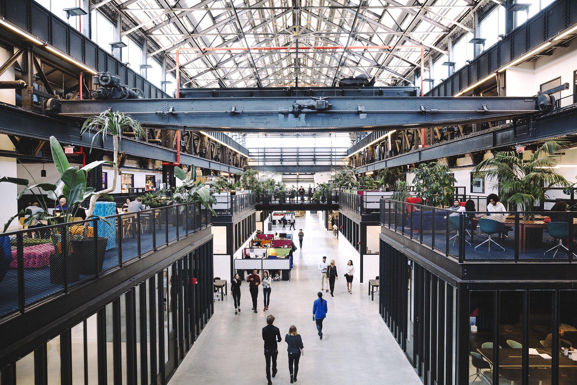 Header image for Urbantech NYC. A spacious industrial-style atrium called New Lab, with people walking and gathering. The space features two levels with black railings, glass partitions, and numerous plants. The ceiling is high and metal beams are visible, creating an open and airy atmosphere. Photo courtesy of New Lab.