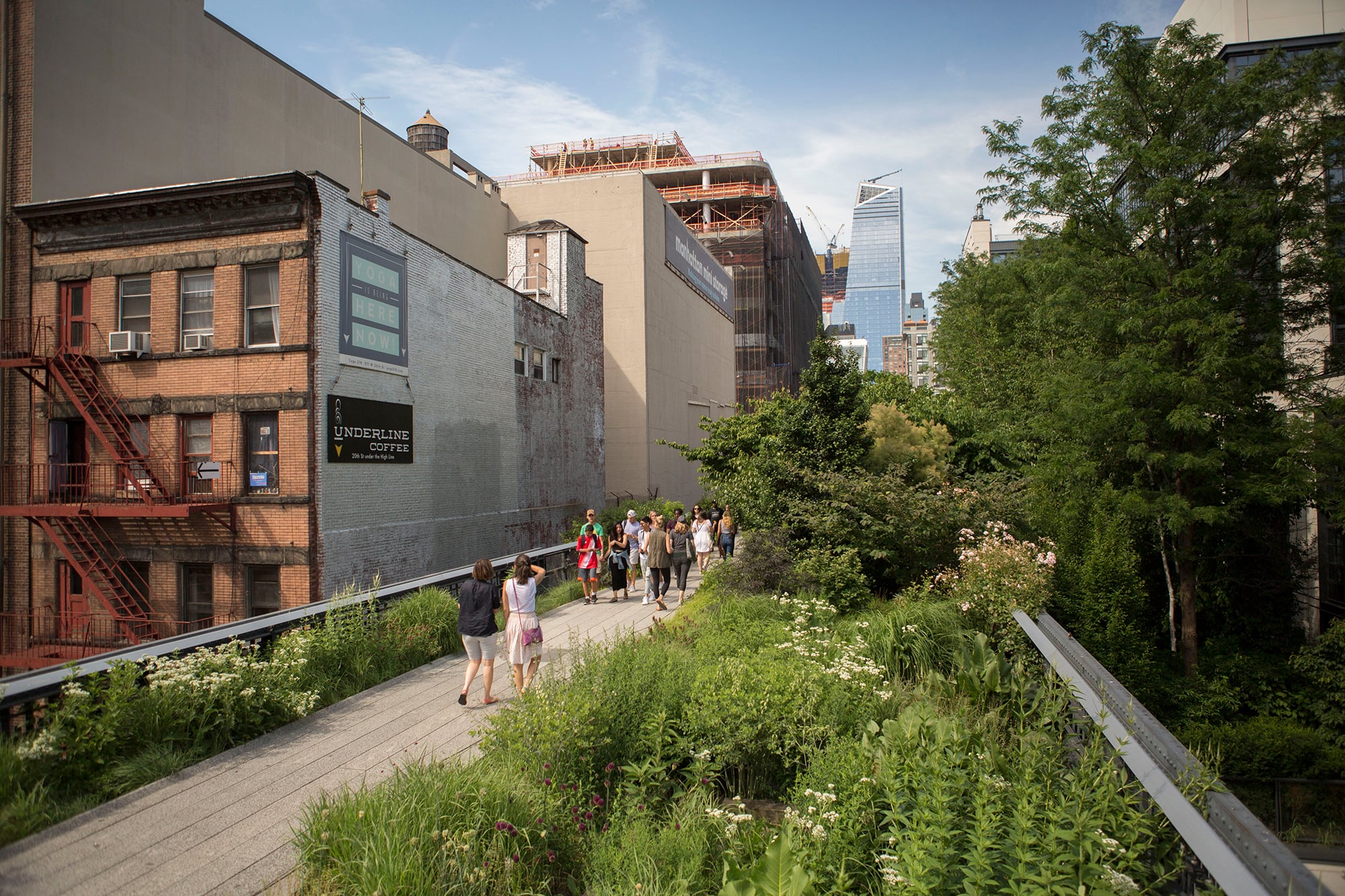 Photo of The High Line by Brittany Petronella. People walk along a landscaped elevated park surrounded by lush greenery, with Manhattan buildings and skyscrapers in the background under a partly cloudy sky.