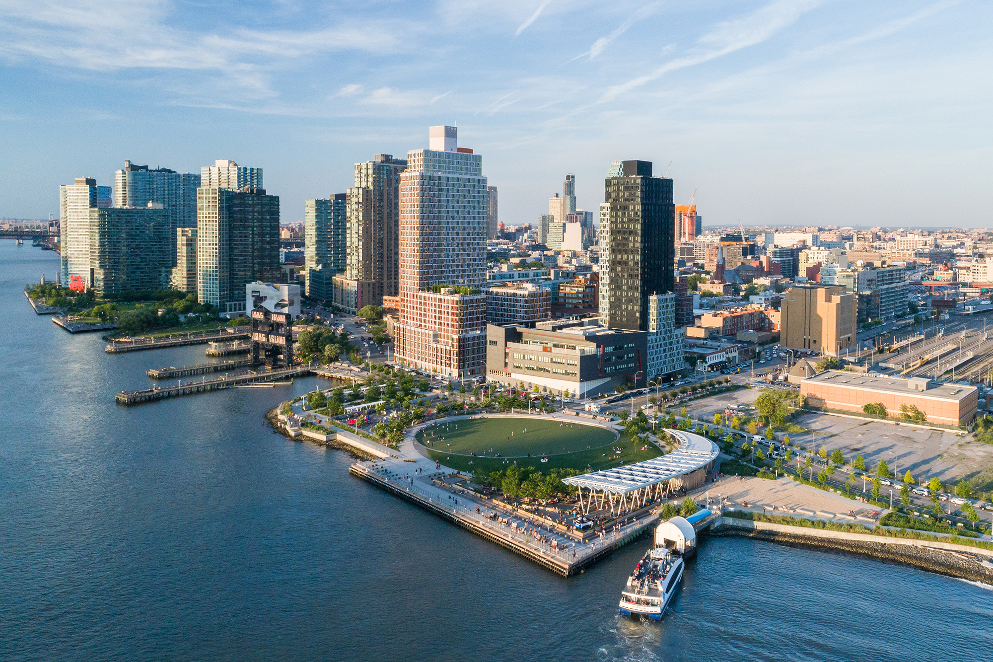 Aerial view of Hunter's Point South in Long Island City, Queens. The park features a large, circular green space surrounded by trees, a ferry terminal with a boat docked, and the East River with high-rise buildings lining the waterfront. Photo by Jonnu Singleton/SWA Group.