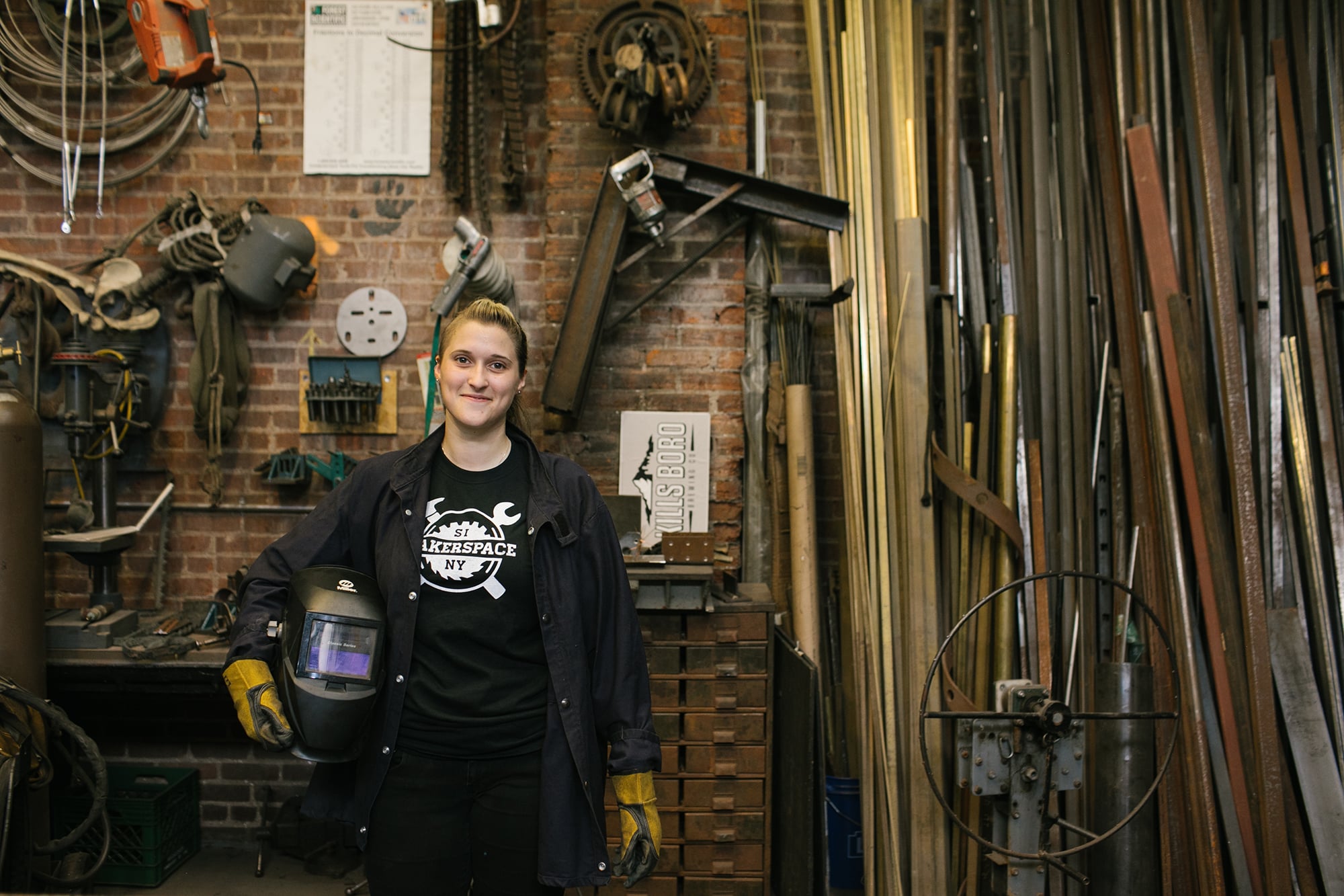 Woman in a Futureworks Makerspace workshop wearing a dark shirt and holding a welding mask. She stands among various tools and materials, including pipes and gears. The wall behind her is brick with hanging tools and gauges.