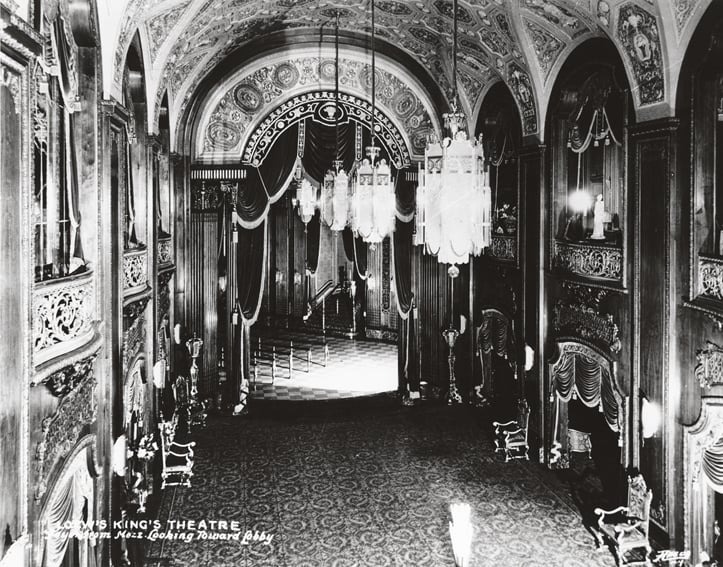 Black-and-white image of a grand theater interior with ornate architecture. Features arched ceilings with intricate designs, large chandeliers, and elaborate curtains around the stage area. Rows of classic seats line the theater, enhancing the opulent atmosphere.