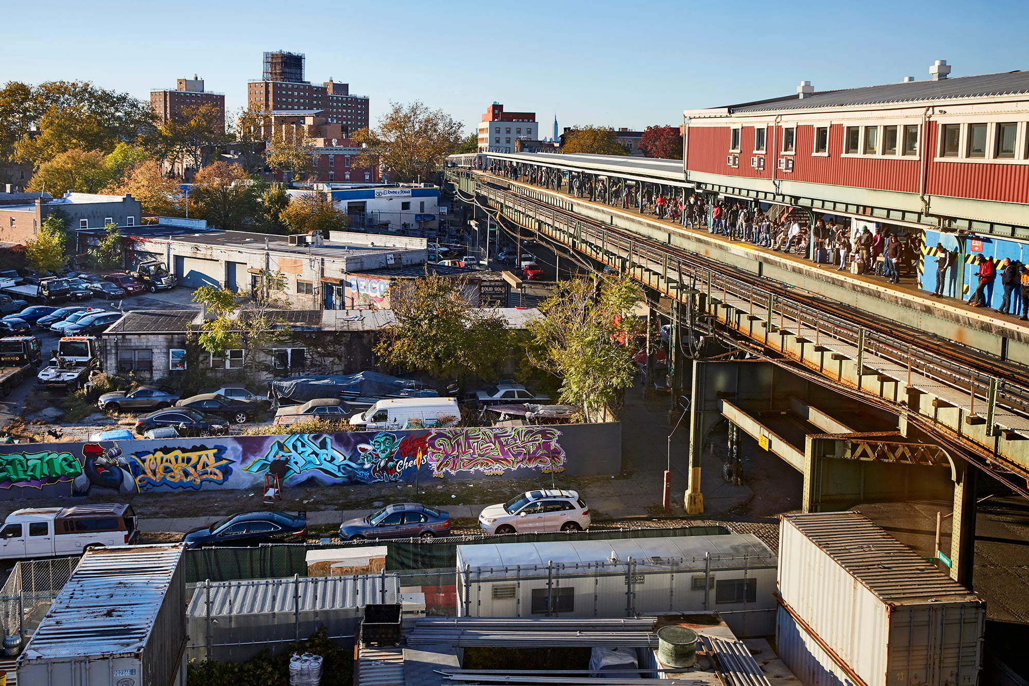Elevated train platform filled with people runs alongside Broadway Junction, lined with parked cars and colorful graffiti-covered walls. Buildings and trees are visible in the background under a clear blue sky. Photo by John Muggenborg/NYCEDC.