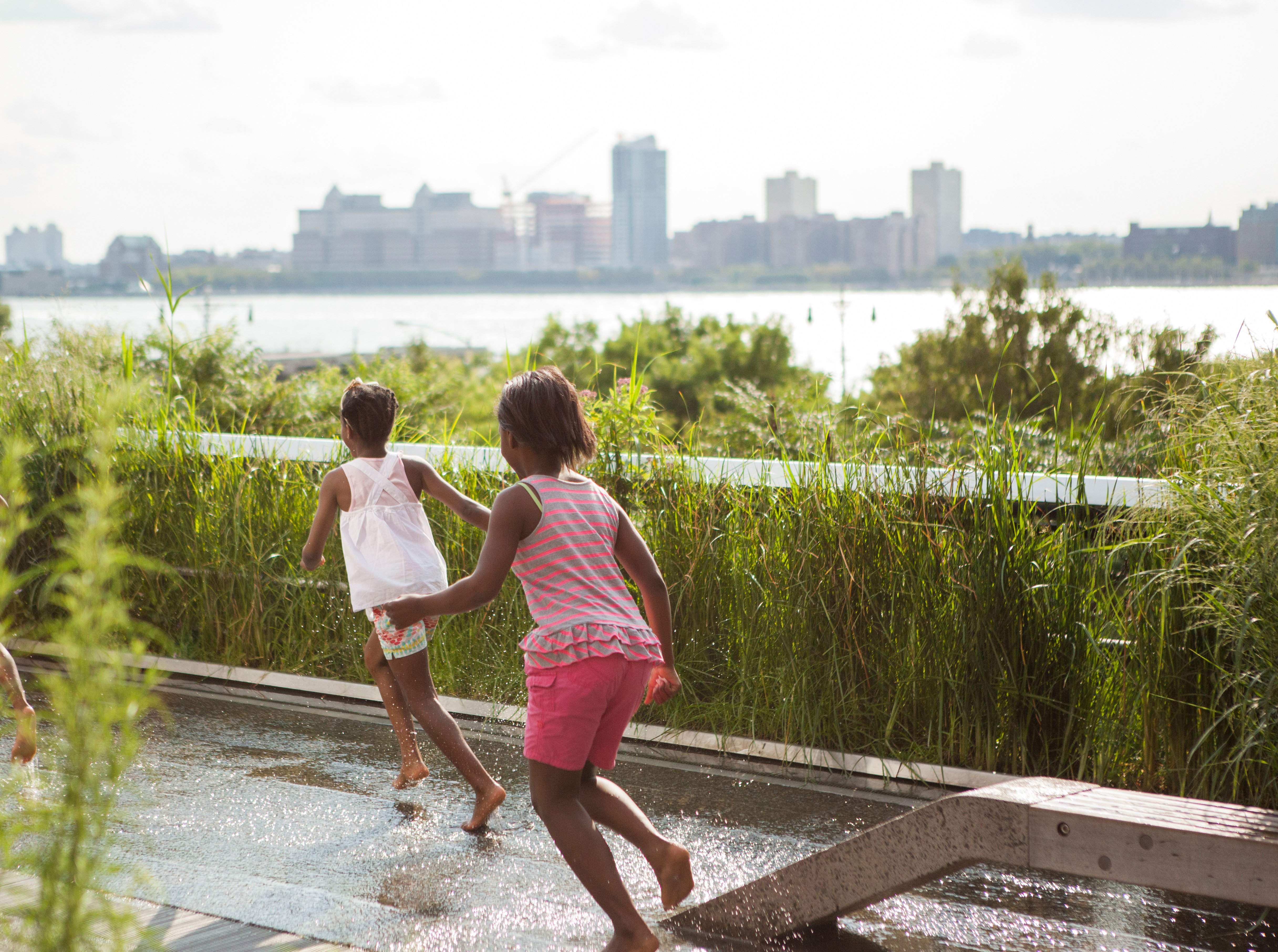 Photo of The High Line by Tagger Yancey IV. Two young children run barefoot through a shallow water feature surrounded by tall grass, with New York skyline and Hudson River visible in the background on a sunny day.