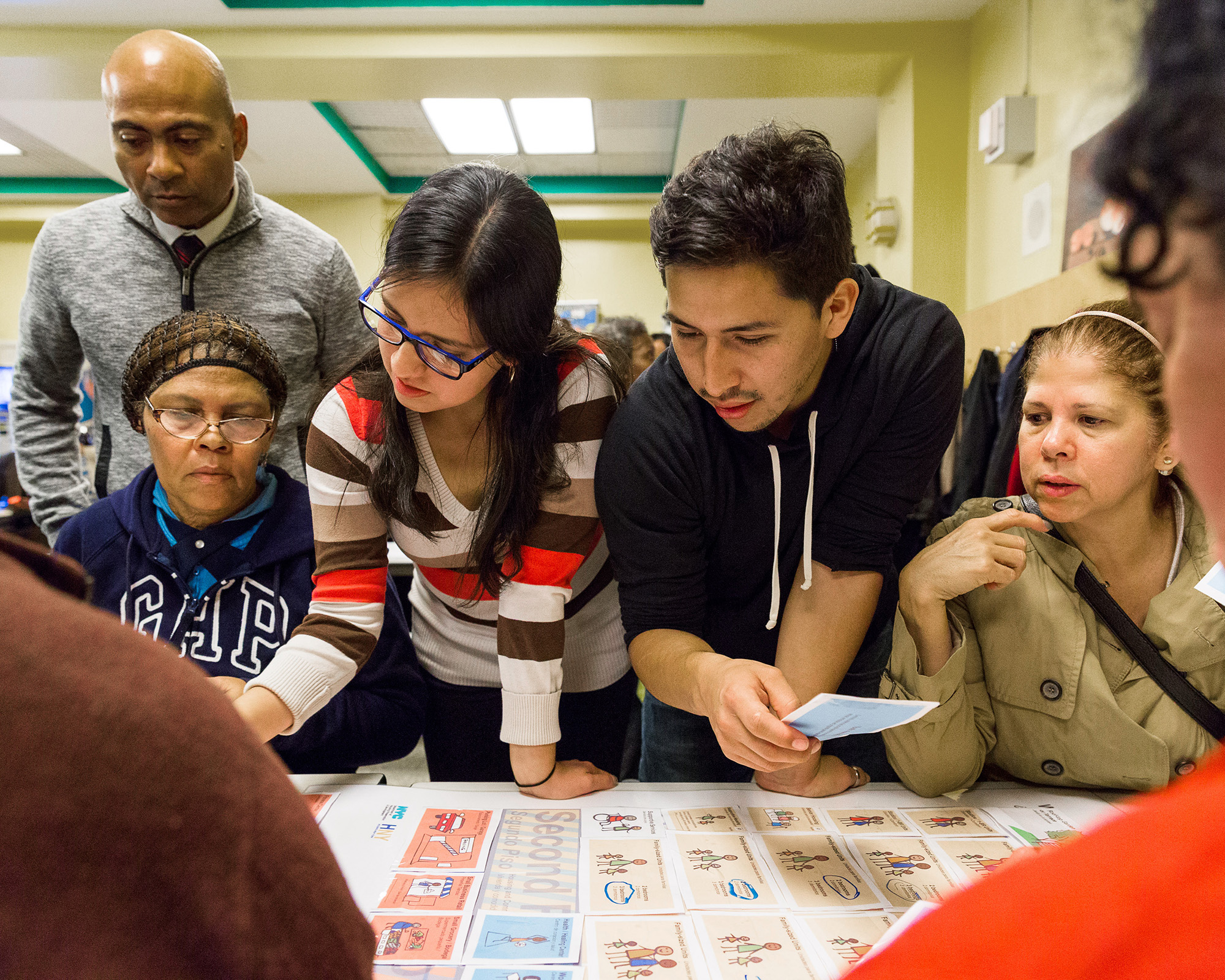 A diverse group of people gathers around a table, intently discussing and pointing at a large sheet with images and text. Some hold papers, suggesting active engagement in a collaborative activity or discussion. The setting appears to be indoors.