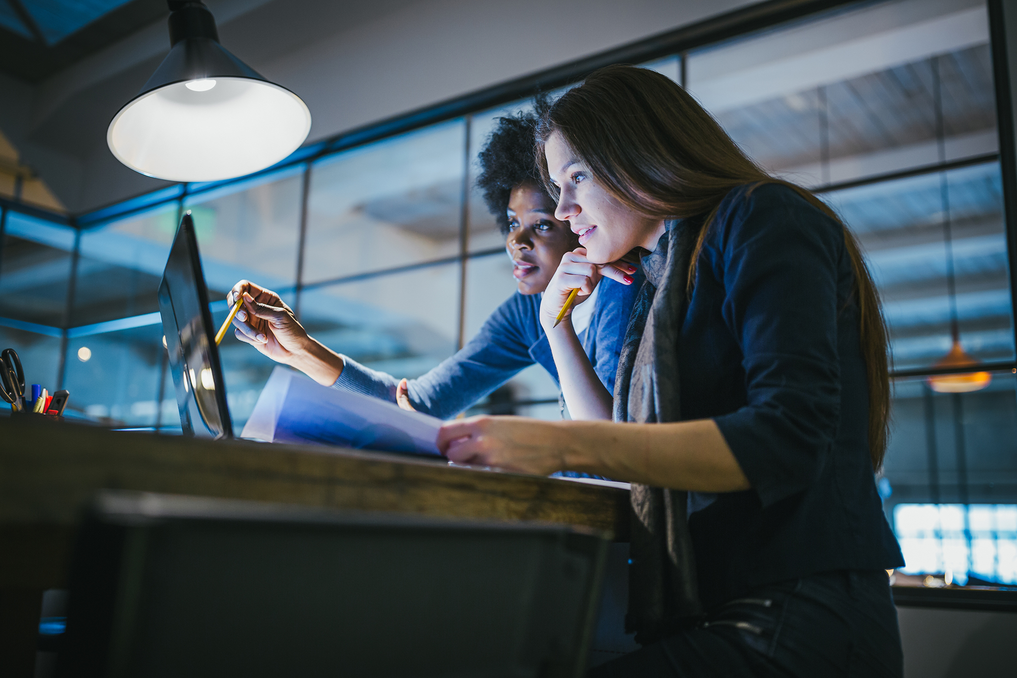 Two women sitting at a desk in a modern office, looking intently at a laptop screen. One holds a pen and points at the screen, while the other holds documents. They appear focused and engaged in their work.