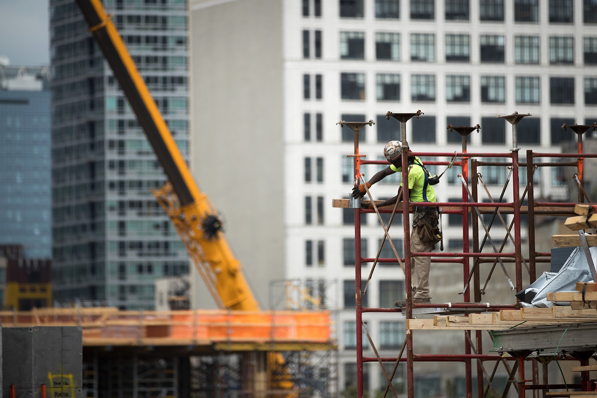 Construction worker wearing a safety harness and helmet stands on metal scaffolding, working with wood planks at a building site in NYC. Tall buildings and a yellow crane are visible in the blurred background. Photo by Drew Angerer/Getty Images.