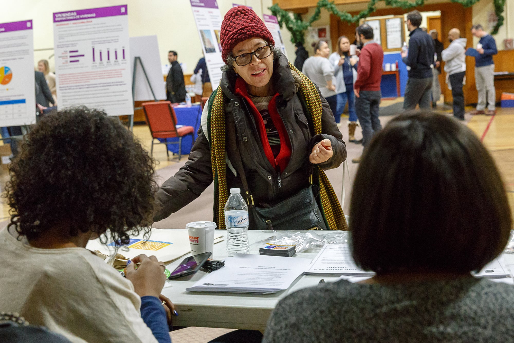 A woman in a red knit hat and scarf talks to two seated individuals at a table in a busy Inwood Spanish Fair, with informational posters and people in the background. The table has papers, a water bottle, and a coffee cup. Photo by Kreg Holt/NYCEDC.