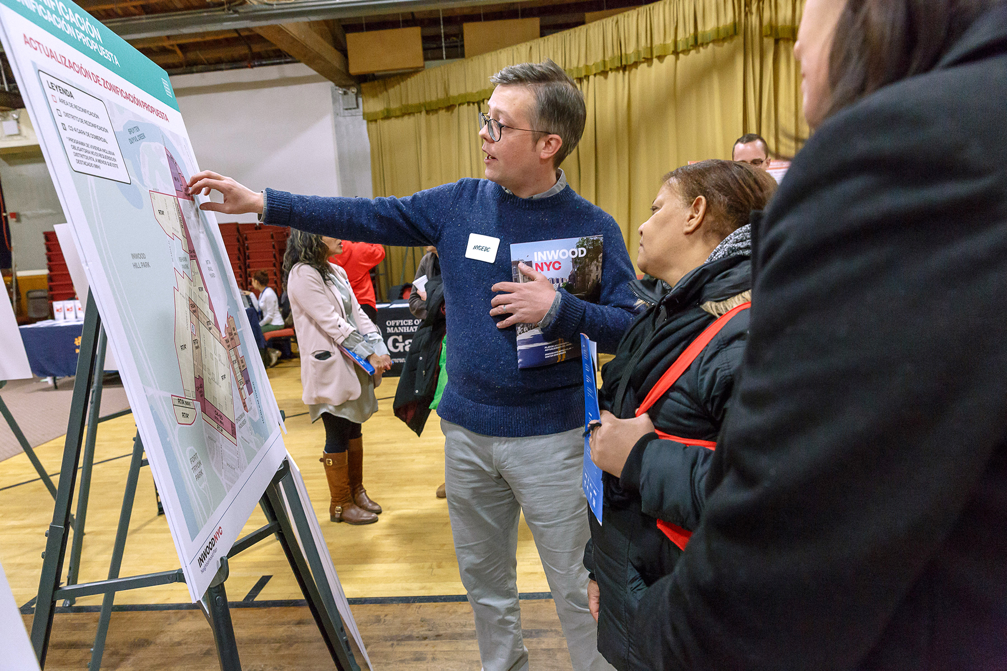 A man wearing glasses and a name badge at Inwood Spanish Fair points at a large map on an easel while speaking to a woman. Other people and informational booths are in the background. Photo by Kreg Holt/NYCEDC.