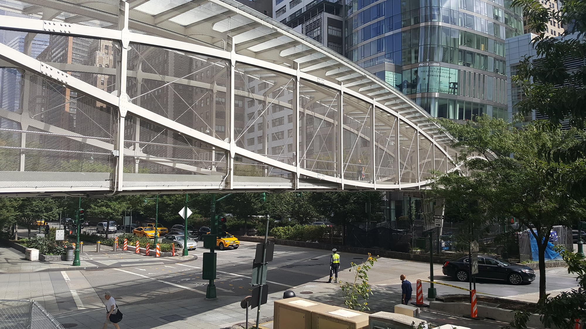 Photo of Robert R. Douglass Bridge by Eric Goodnight/NYCEDC. A white pedestrian bridge with a metal mesh crosses over an NYC street lined with modern glass buildings, trees, taxis, and people, some of whom appear to be construction workers in safety vests.