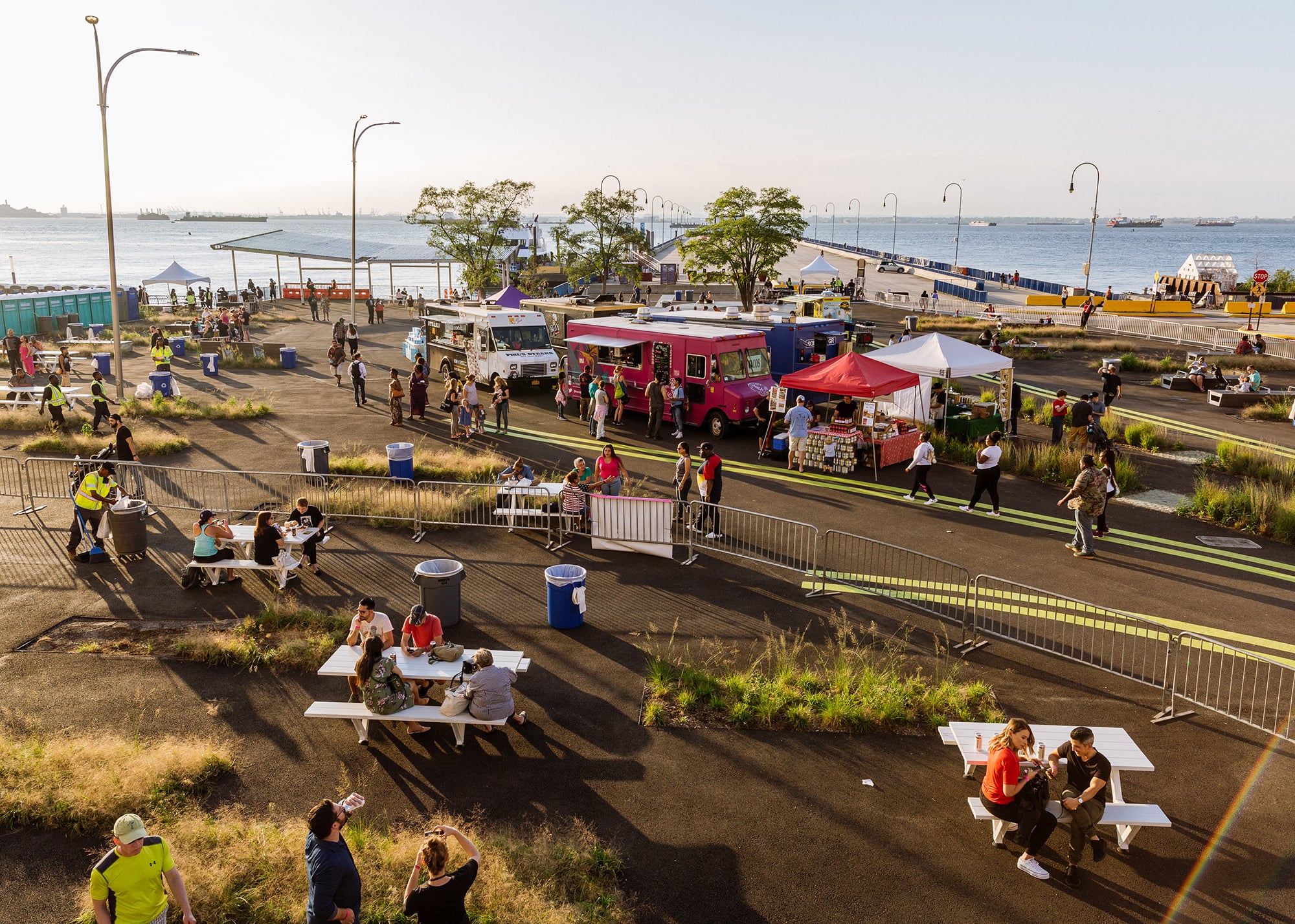 Aerial view of BAT Block Party, showing a waterfront food festival featuring food trucks, vendor tents, and picnic tables. People are enjoying food and mingling. The scene is set against a backdrop of water and a clear sky, with ships visible in the distance. Photo by Kreg Holt/NYCEDC.