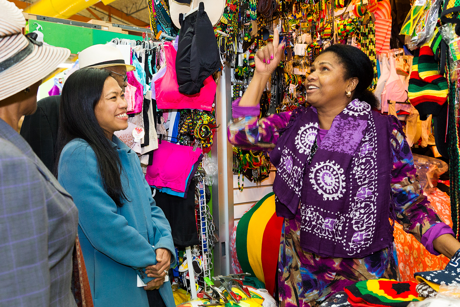 A woman in a purple outfit animatedly gestures while talking to a smiling woman in a blue coat at a colorful Flatbush Caton Market stall selling swimwear and accessories. Other shoppers are partially visible.