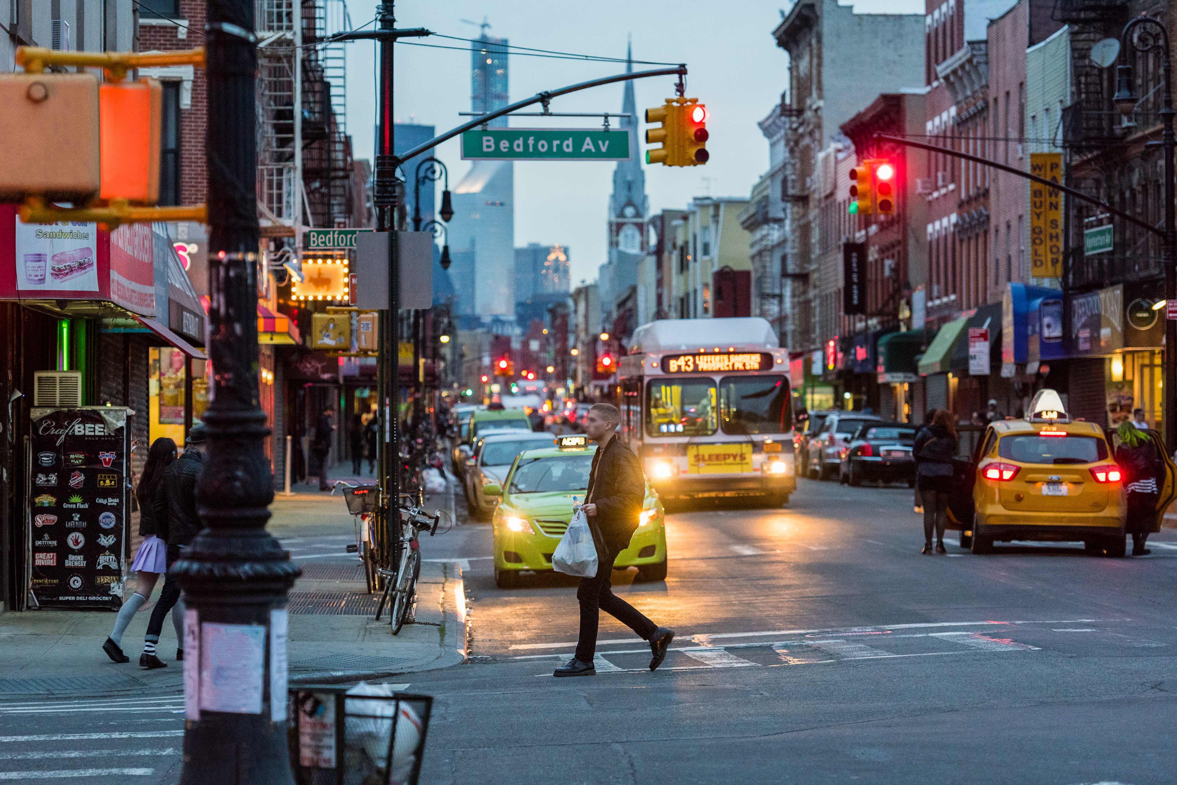 Busy NYC street scene at dusk shows people crossing the road, yellow taxis, a bus, shops, bicycles, and bright traffic lights under a sign reading Bedford Av. Tall buildings are visible in the background. Photo by Jen Davis.