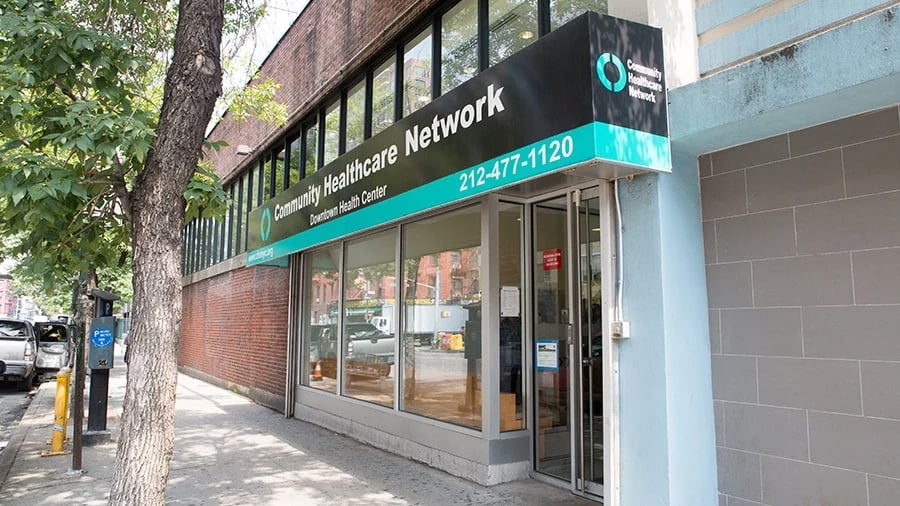 A street view of the CHN East New York health clinic, showing its glass entrance, a teal and black sign with the centers name and phone number, and a tree lining the sidewalk outside.
