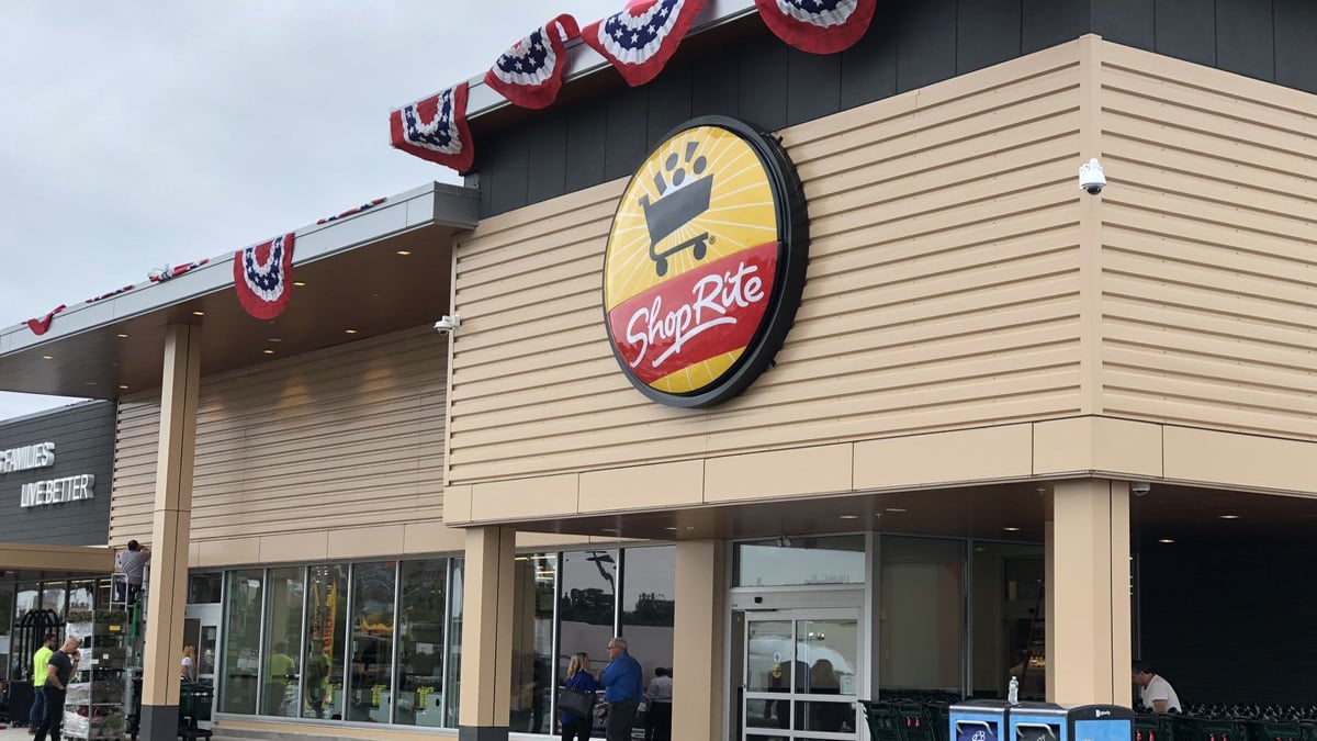 The exterior of a ShopRite store in Soundview, Bronx, with a large round ShopRite logo sign above the entrance. Red, white, and blue bunting decorates the roof edge. Several people are near the entrance.
