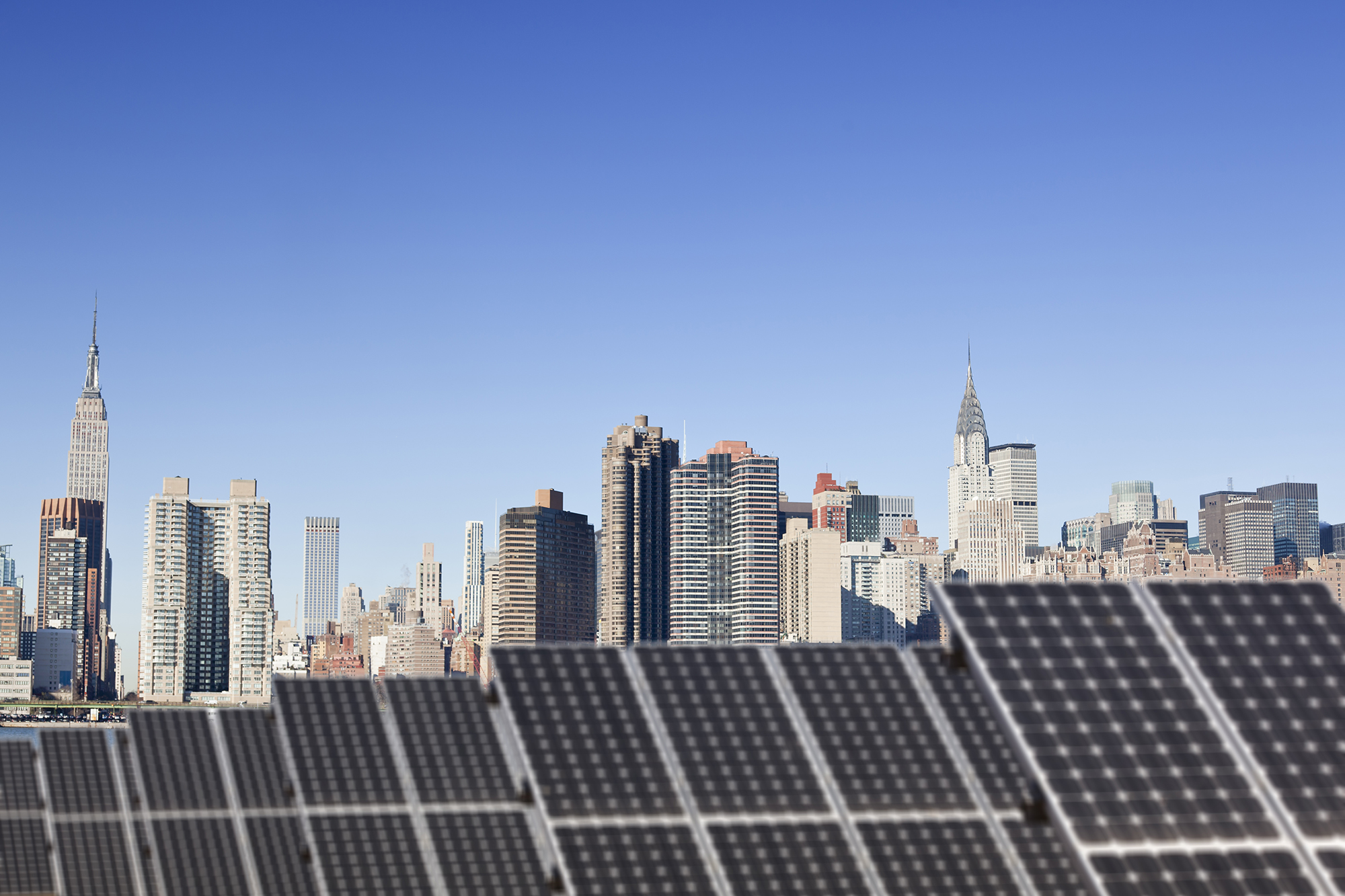 Rows of solar panels in the foreground with a clear blue sky and the New York City skyline, including the Empire State Building and Chrysler Building, in the background.