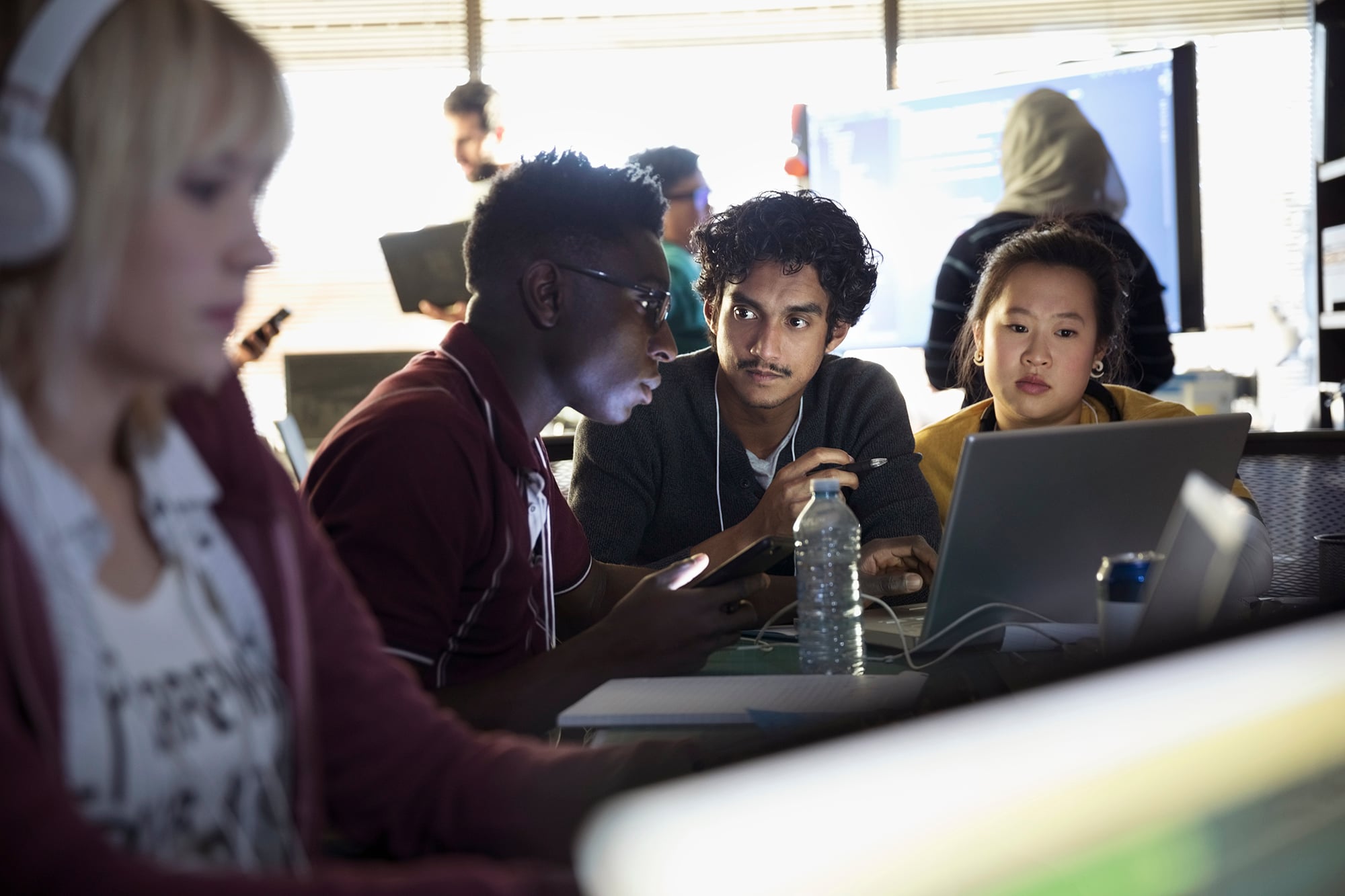 Three people of color sit at a table, focused on a laptop screen. One person holds a phone, while others listen attentively. The background shows more people collaborating in a bright, busy tech office setting.
