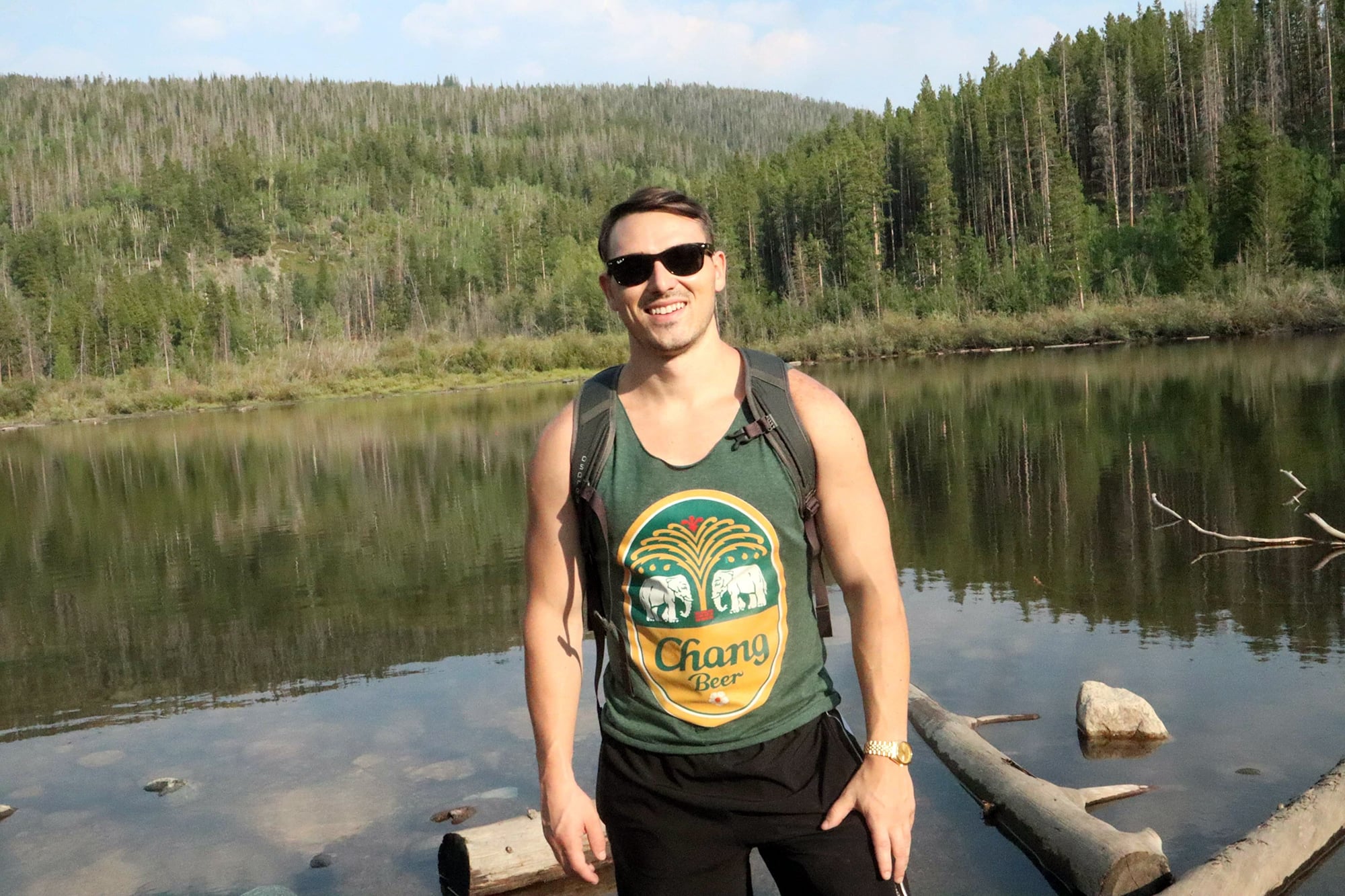 Daniel Clark smiling while wearing sunglasses and a tank top stands in front of a serene lake surrounded by pine trees. They have a backpack on and are standing on a rocky shore, with forested hills and a blue sky in the background.