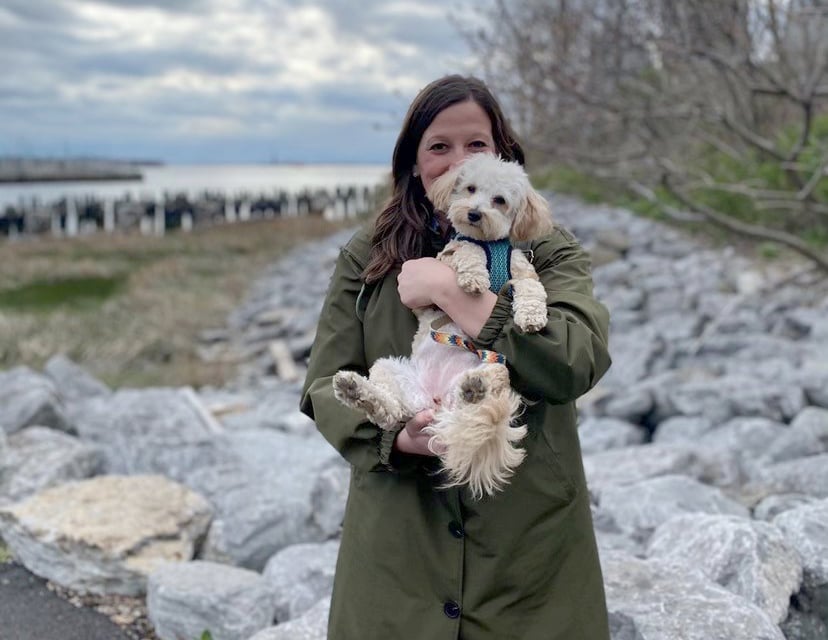 Emily De Vito in a dark green coat stands on a rocky shoreline, holding her small, fluffy white dog. The sky is cloudy, and theres water and some distant piers in the background.