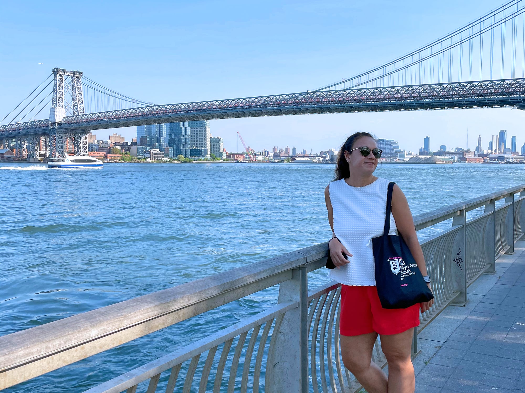 Jennifer Montalvo in sunglasses leans on a railing by the waterfront, wearing a white top and red skirt, with a tote bag. Manhattan Bridge is in the background under a clear blue sky.