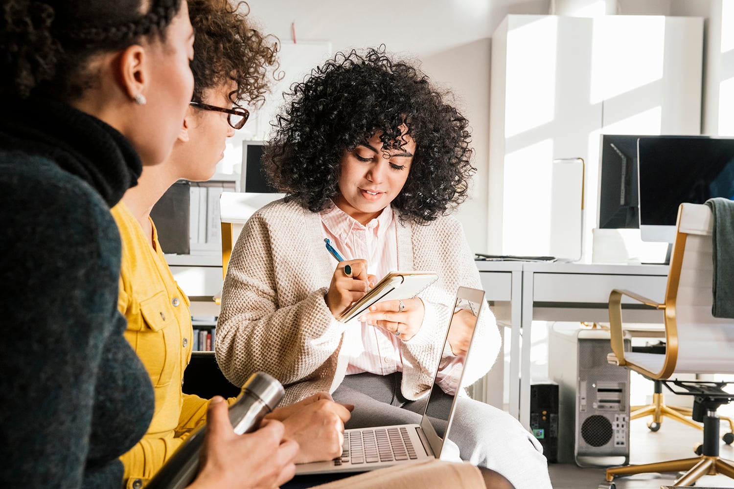 Three women work together in a bright office space. One woman writes in a notebook, while the others listen attentively. A laptop is open on one lap, and office equipment is visible in the background.