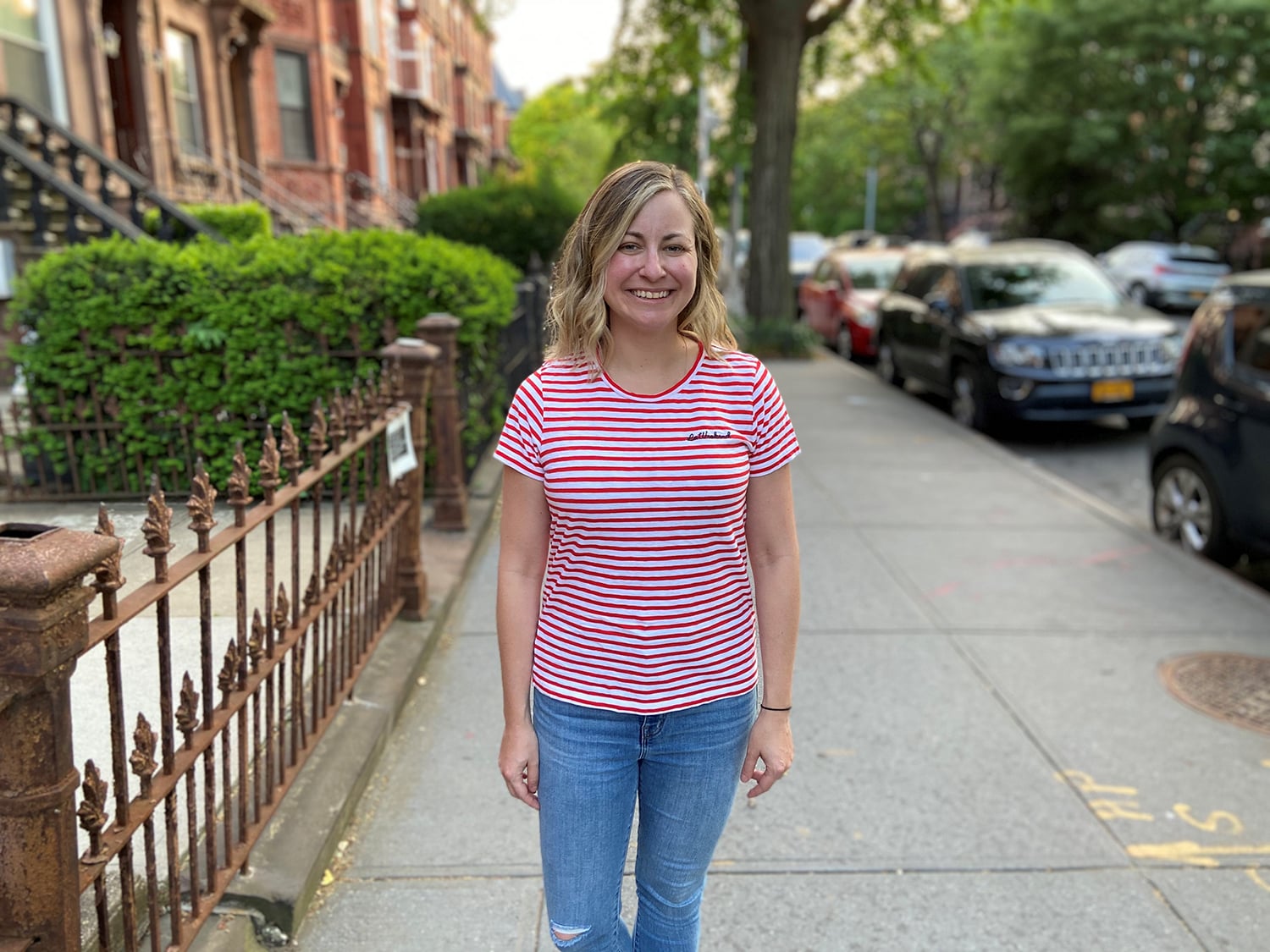 A woman with shoulder-length hair, Melissa Pumphrey, wearing a red and white striped shirt and jeans stands smiling on a sidewalk. There are parked cars and green trees lining the street, and an iron fence is visible on the left.
