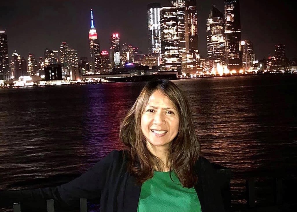 Stella Maniago in a green top and black jacket stands smiling beside a waterfront at night, with a brightly lit city skyline featuring a prominent building with red and blue lights in the background.