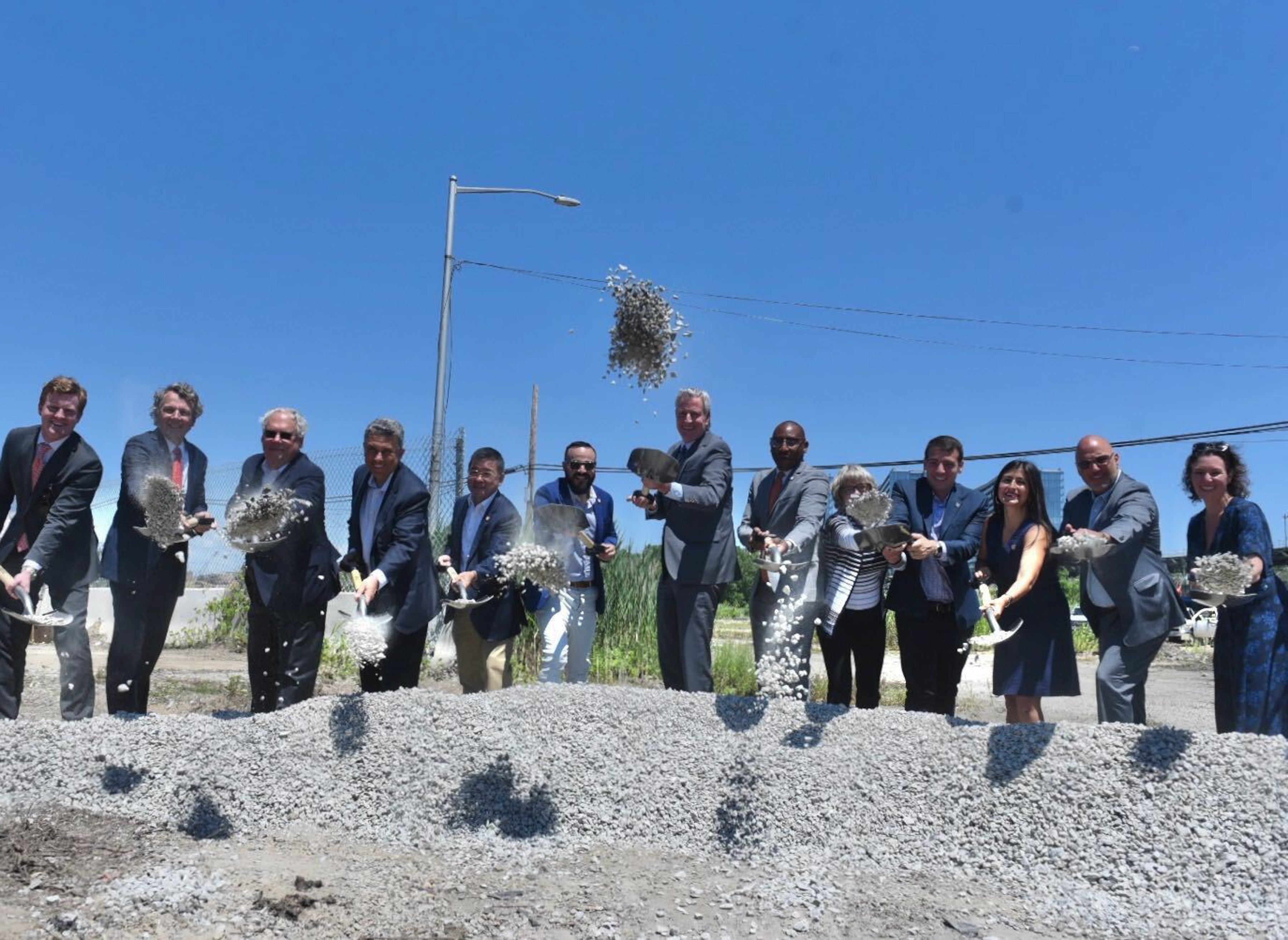 A group of people in business attire participate in Willets Point environmental remediation groundbreaking ceremony, tossing gravel with shovels into the air on a sunny day, standing in front of a construction site.