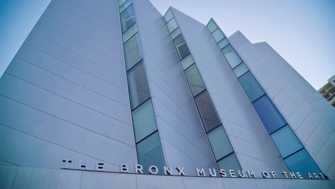 A modern, angular building with tall, narrow windows and the words The Bronx Museum of the Arts displayed in large letters on the facade. The photo is taken from a low angle against a clear sky. Photo by Ian Spanier.