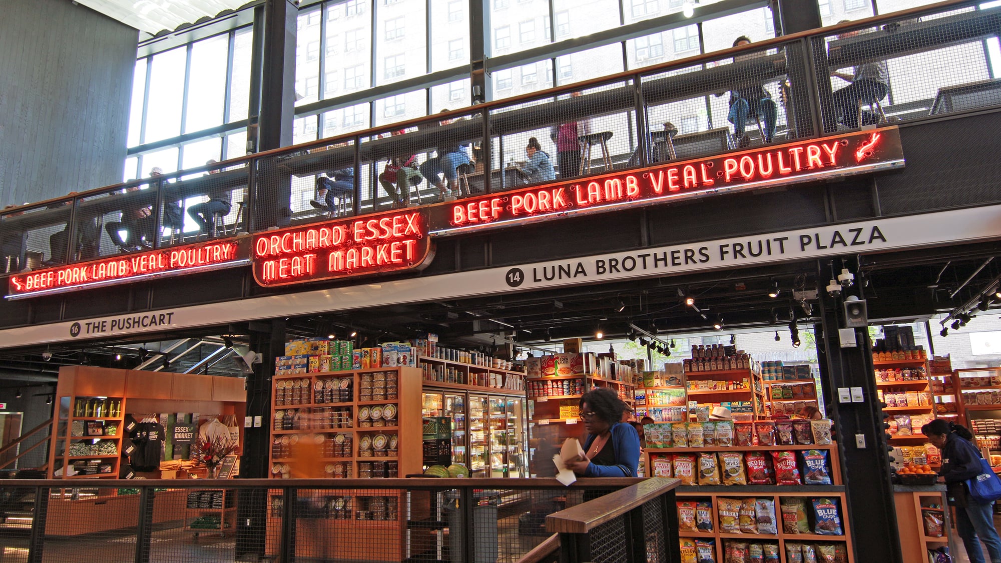 A spacious indoor market with shelves of groceries, neon signs reading “BEEF PORK LAMB VEAL POULTRY” and “ORCHARD ESSEX MEAT MARKET,” and people shopping or sitting on an upper level with large windows.
