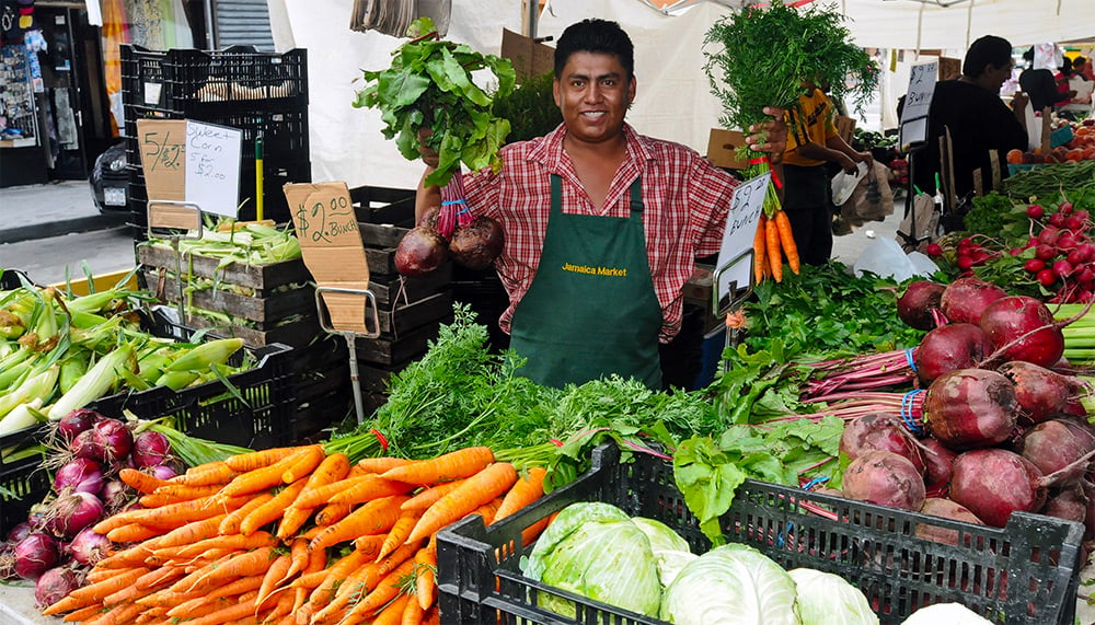 A smiling vendor in a green apron stands behind a market stall displaying carrots, beets, onions, lettuce, and other fresh vegetables, holding bunches of carrots and beets in each hand.