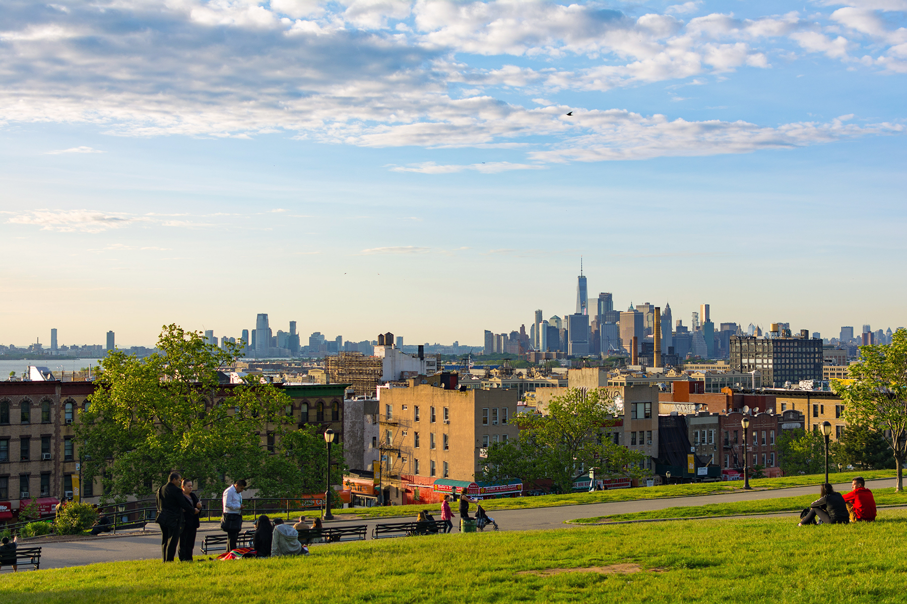 A grassy hill in Sunset Park, Brooklyn overlooking the New York City skyline at sunset. People sit on the grass and benches, enjoying the view. The sky is partly cloudy, with the sun casting warm light on the scene. Tall buildings are visible in the distance.