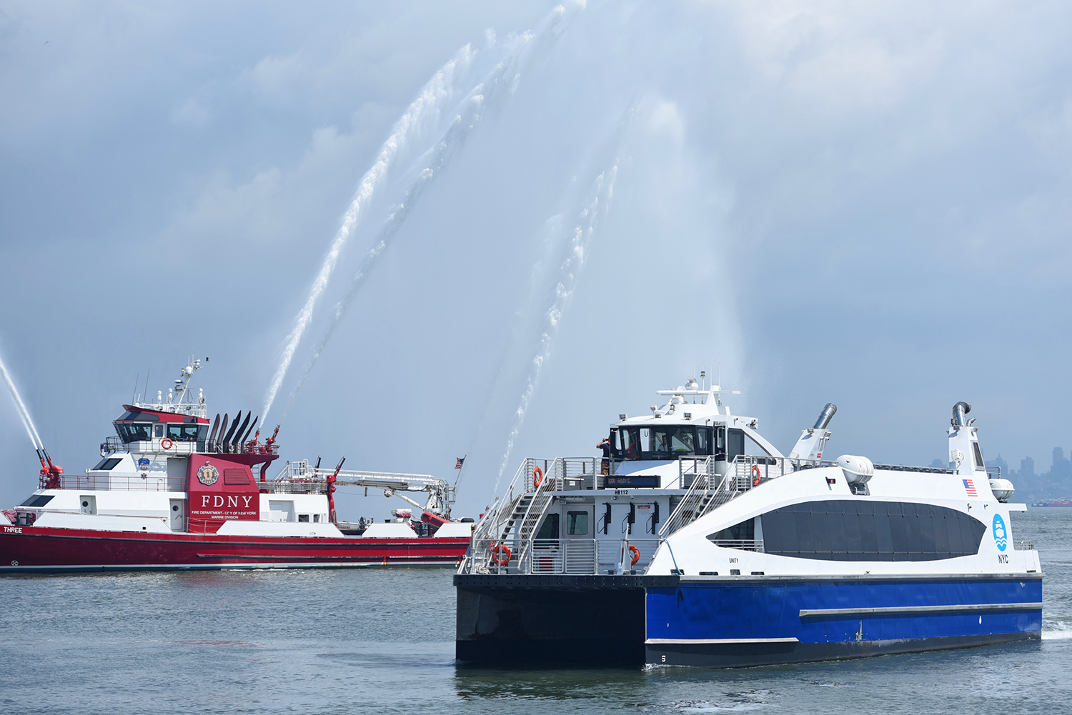 A red FDNY fireboat sprays water jets into the air near a white and blue St. George NYC Ferry on the water, with a cloudy sky and New York City skyline faintly visible in the background.