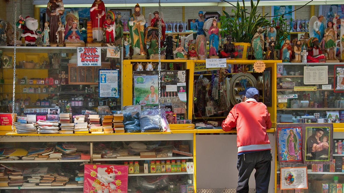 A person in a red jacket stands at a colorful outdoor shop counter filled with religious statues, candles, books, and various spiritual items displayed on shelves and hanging above.