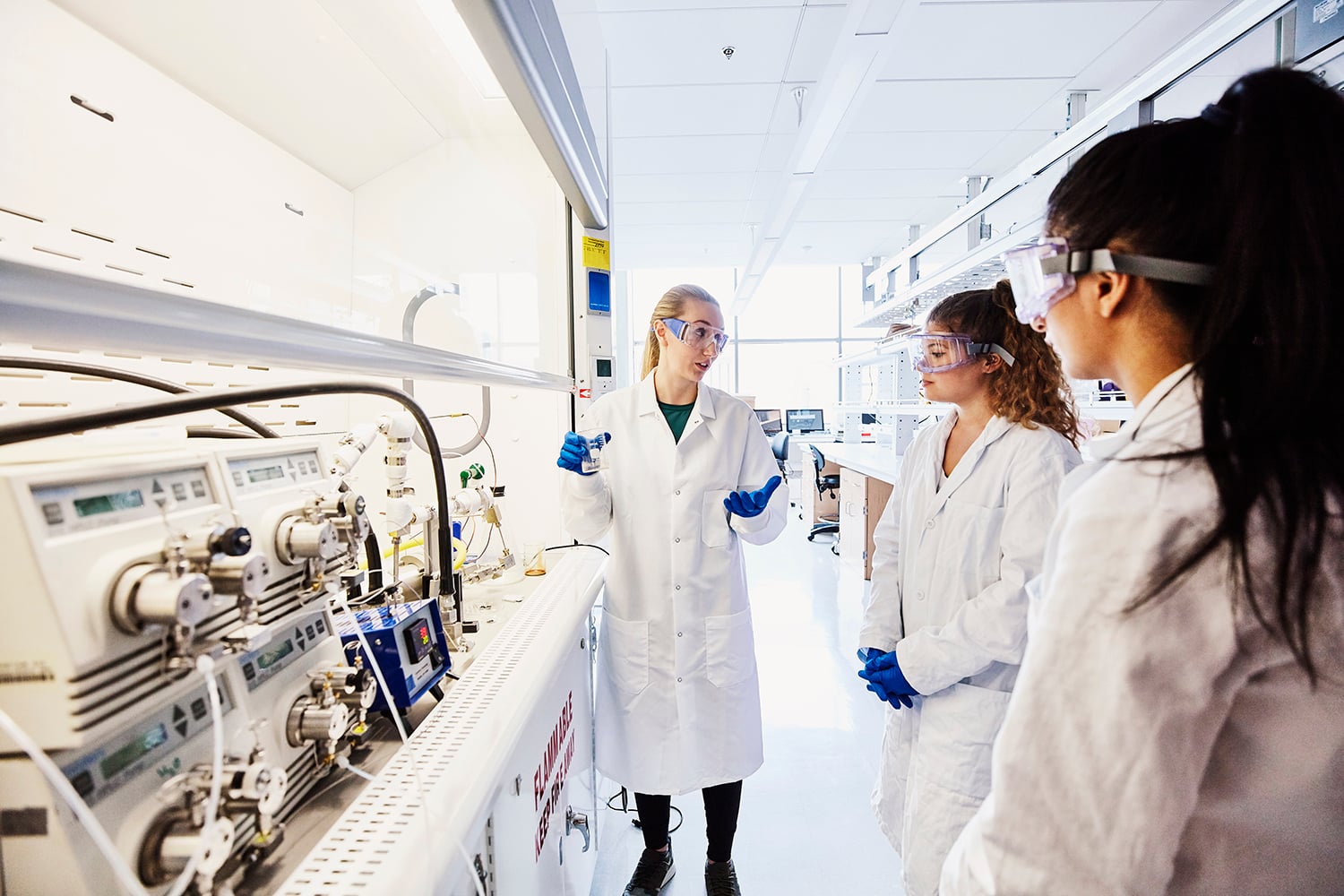 Three scientists wearing lab coats and safety goggles discuss in a bright laboratory next to scientific equipment and machinery. One person gestures while holding a tablet, and the others listen attentively.
