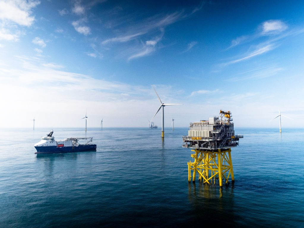 Large offshore wind platform with yellow supports stands in the sea near a service ship. Wind turbines are visible in the distance under a blue sky with light clouds.