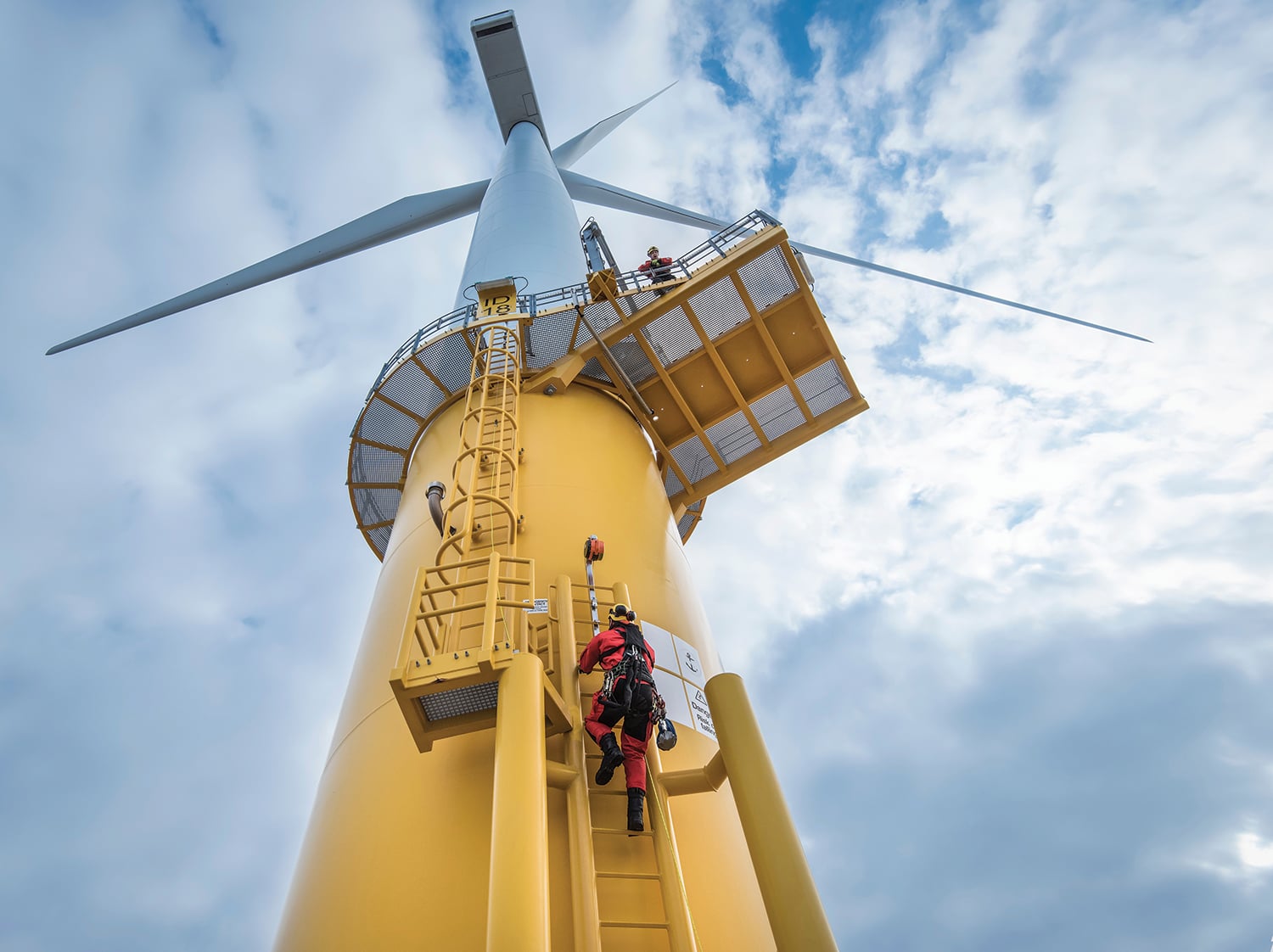 Worker in red safety gear climbs a yellow ladder on a large offshore wind turbine tower. Another person is visible on a platform above. The sky is cloudy, and the turbine blades extend outward from the top.