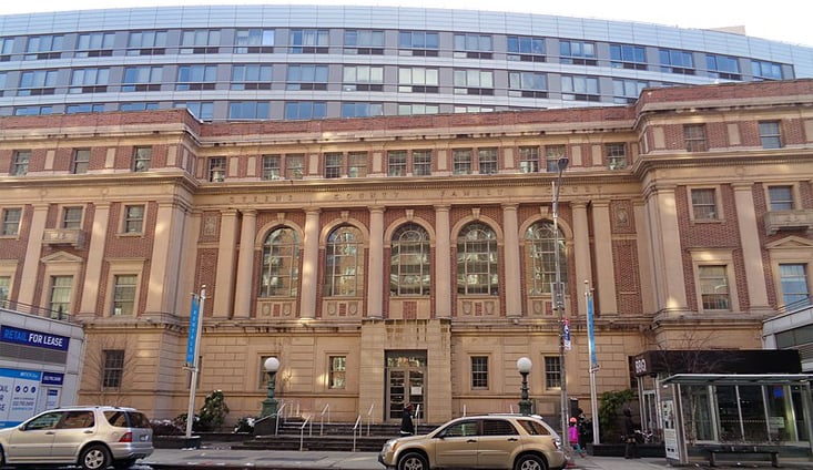 Photo of GJDC Business Training Center shows a historic brick and stone building with large arched windows and columns, set beneath a modern glass building. Cars are parked along the street in front, and blue flags are visible on both sides of the entrance.