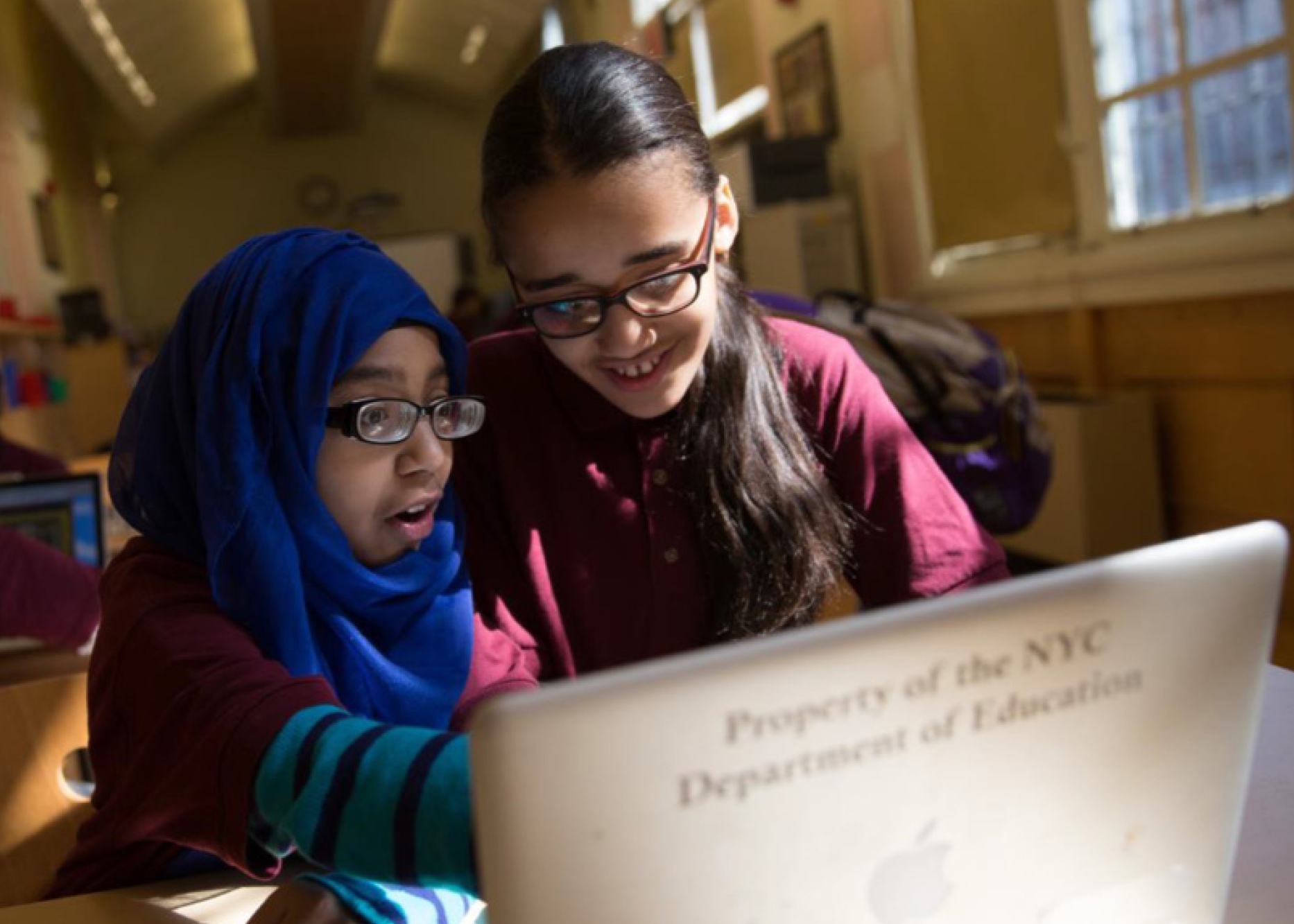 Two female students at P.S. 188, The Island School, one wearing a blue hijab, look excitedly at a laptop with Property of the NYC Department of Education on it. They're in a classroom with sunlight streaming through the windows.