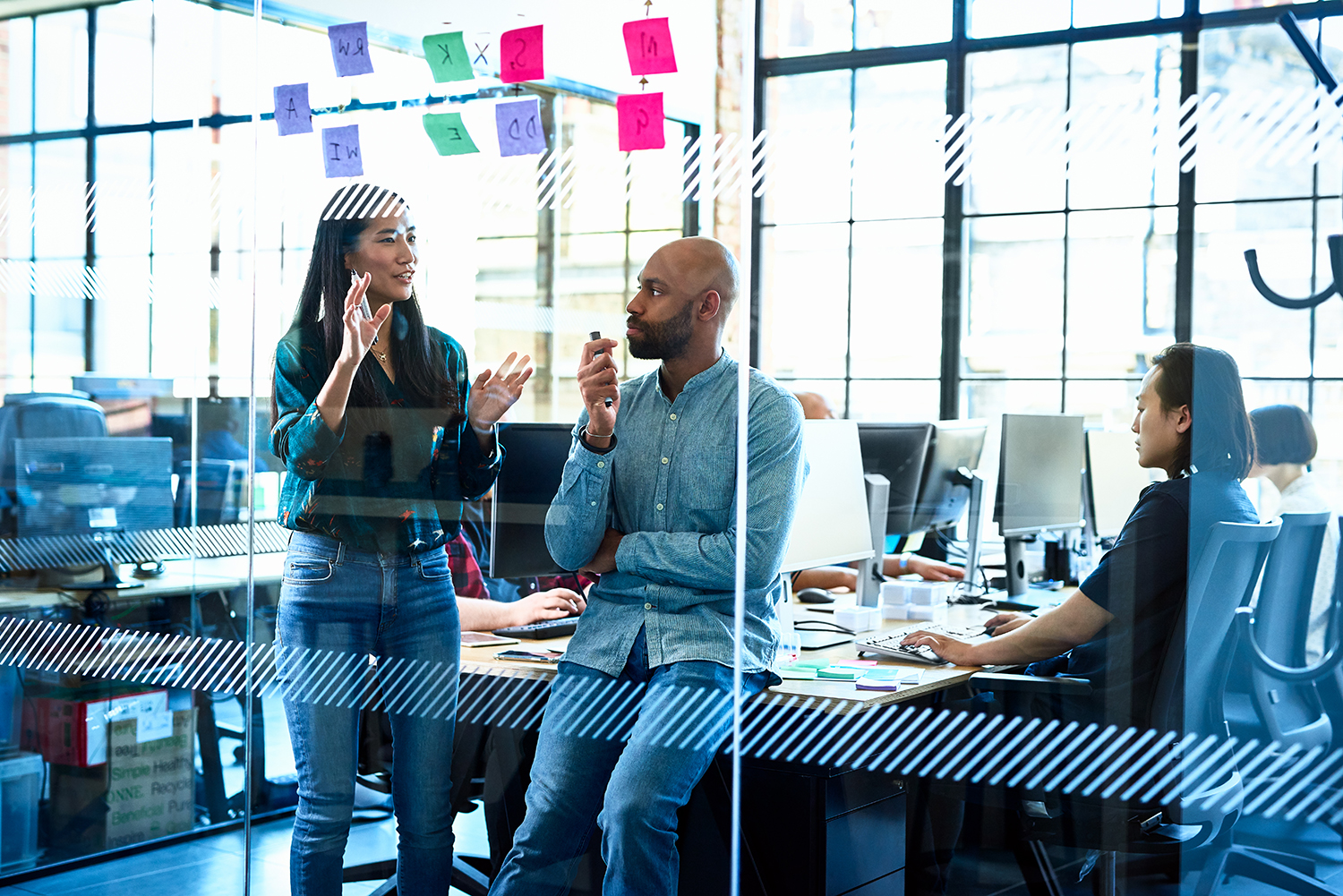 Header image of Venture Access NYC program page. A diverse group of people in a modern office setting. A woman is standing and gesturing while a man sits on a desk, listening. Another person works on a computer in the background. Colorful sticky notes are visible on the glass wall.