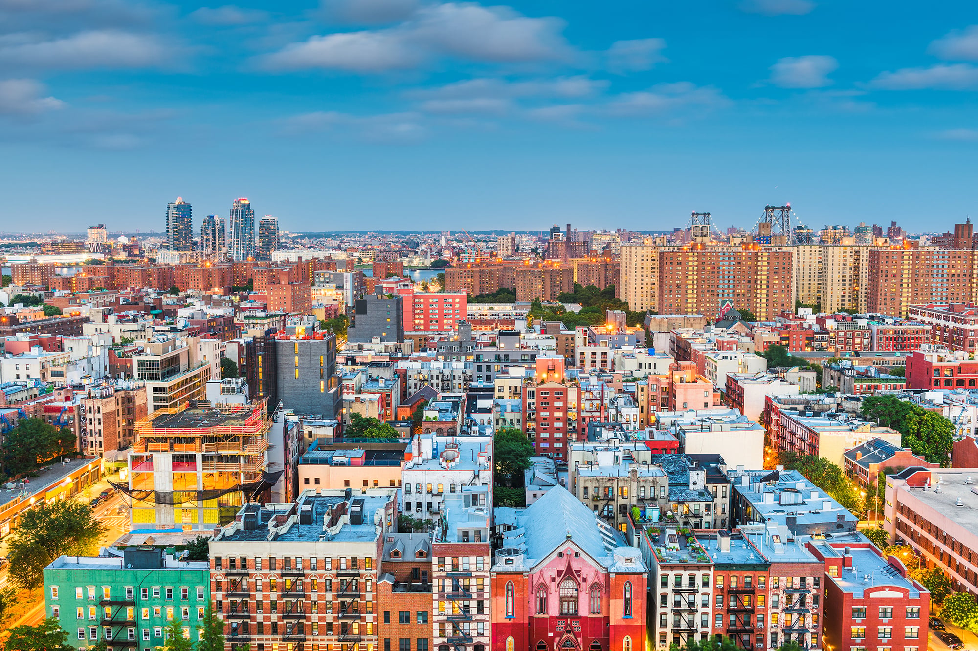 Proptech Piloting Program portrayed through an aerial view of a vibrant New York City at dusk with colorful buildings, some under construction, and a prominent red-roofed church. High-rise apartments and distant bridges are visible under a partly cloudy sky.