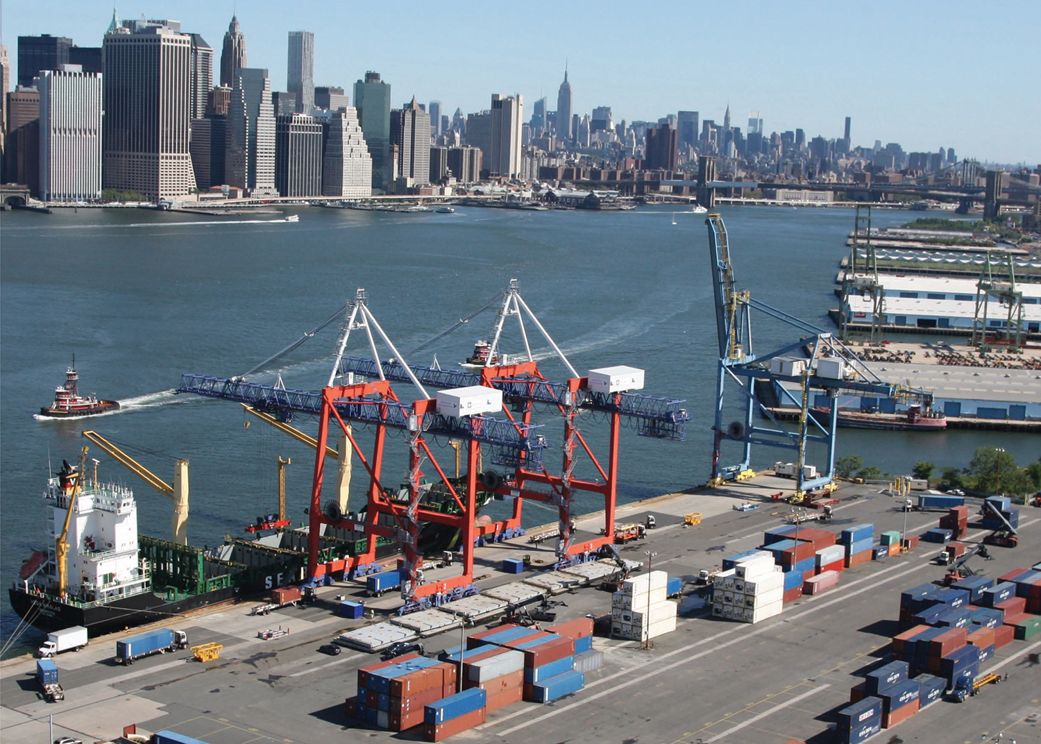 Busy port with colorful shipping containers, cranes loading a cargo ship, and tugboats, set against New York City skyline with tall buildings across the river under a clear sky.