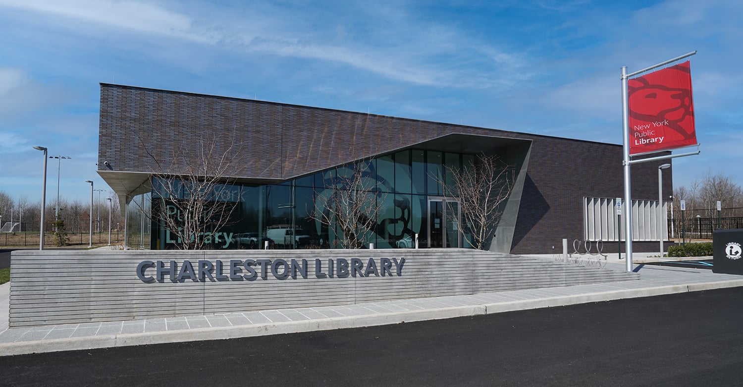 Modern building with dark brick exterior labeled “Charleston Library.” Large windows show part of the New York Public Library lion logo inside. A red NYPL flag hangs on a pole outside; clear blue sky overhead.