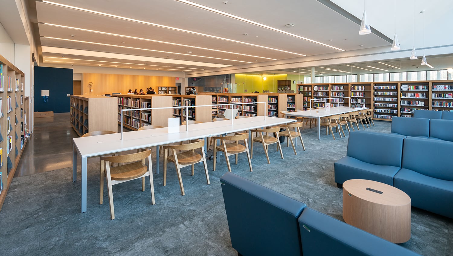 Spacious and modern Charleston Library with long white tables, wooden chairs, blue cushioned seating, and rows of bookshelves. Pendant lights hang from the ceiling, and a few people are visible at the far end of the room.