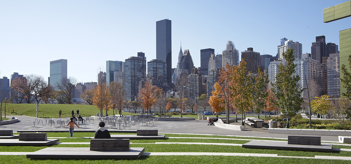 People sit on benches in Cornell Tech Campus with green lawns and autumn trees, with a clear view of the New York City skyline and skyscrapers under a clear blue sky.