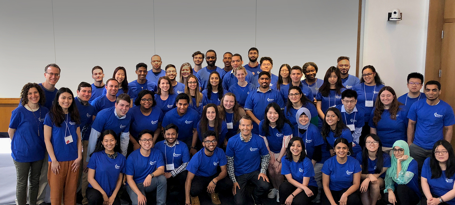 LifeSci NYC interns wearing matching blue shirts pose together indoors, smiling at the camera. The background is a plain white wall with a camera mounted in the top right corner.